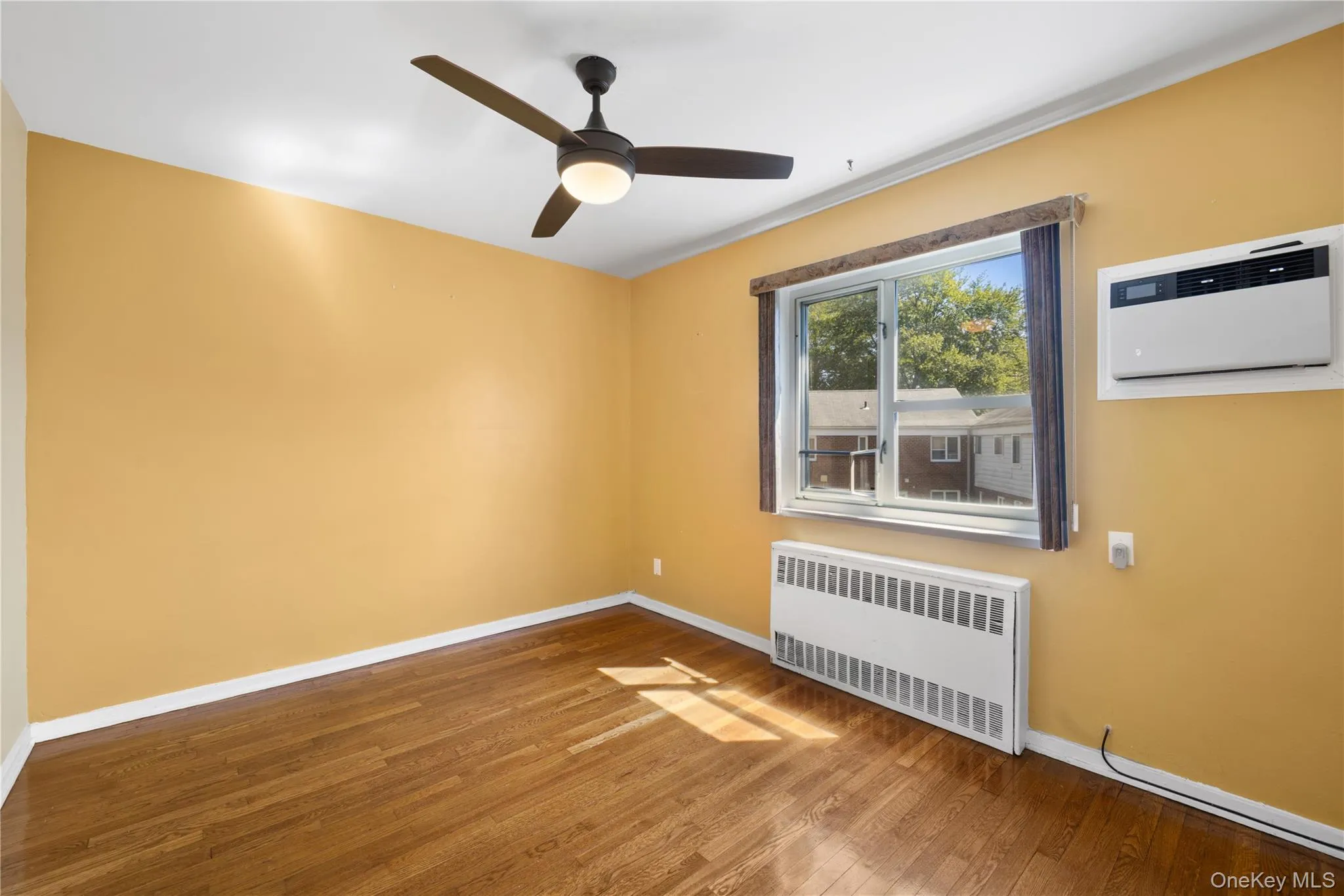 Empty room featuring radiator, wood finished floors, a wall mounted air conditioner, and a ceiling fan Empty room featuring radiator, wood finished floors, a wall mounted air conditioner, and a ceiling fan