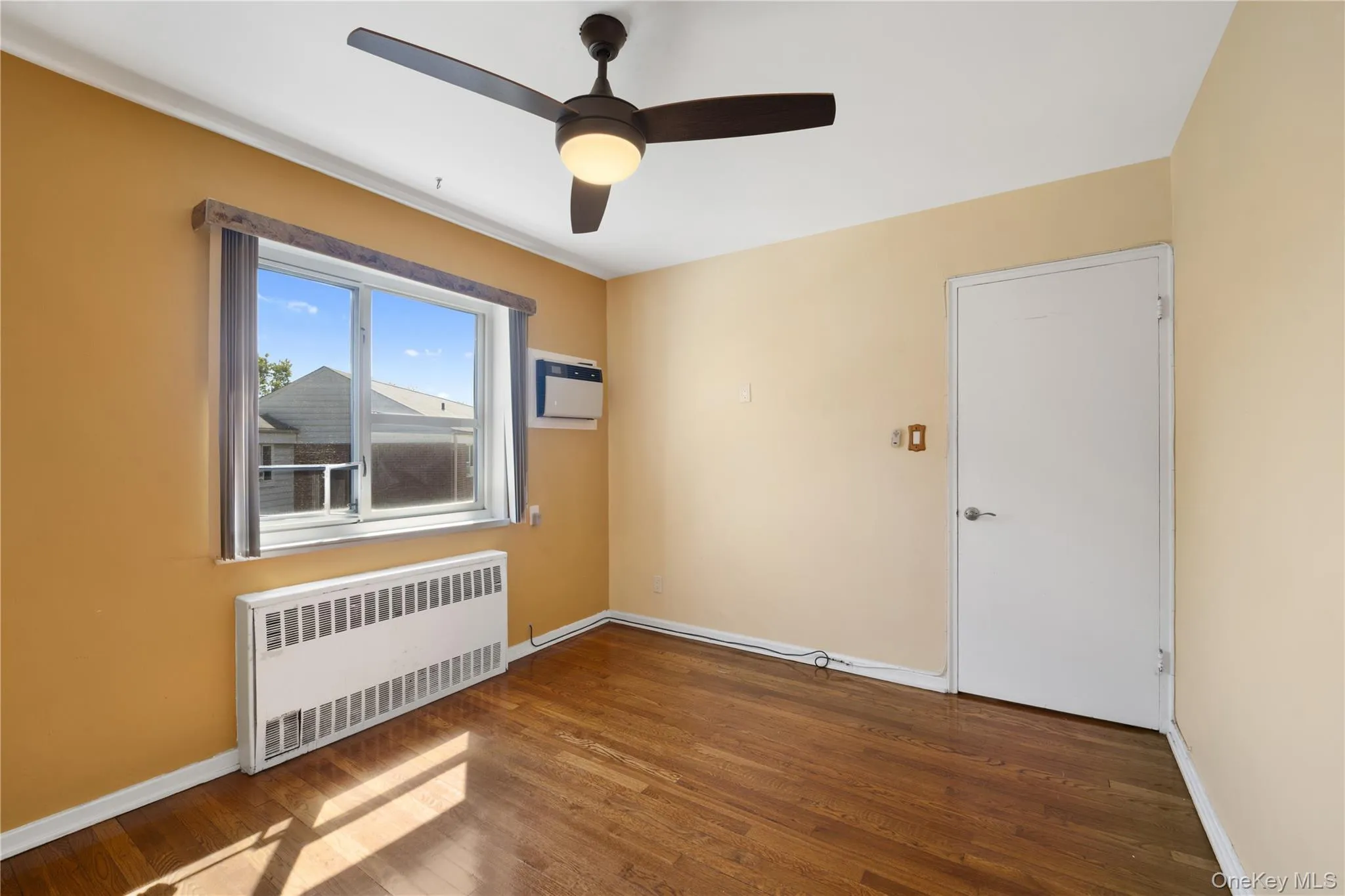Empty room with radiator, dark wood-type flooring, a ceiling fan, and a wall mounted AC Empty room with radiator, dark wood-type flooring, a ceiling fan, and a wall mounted AC