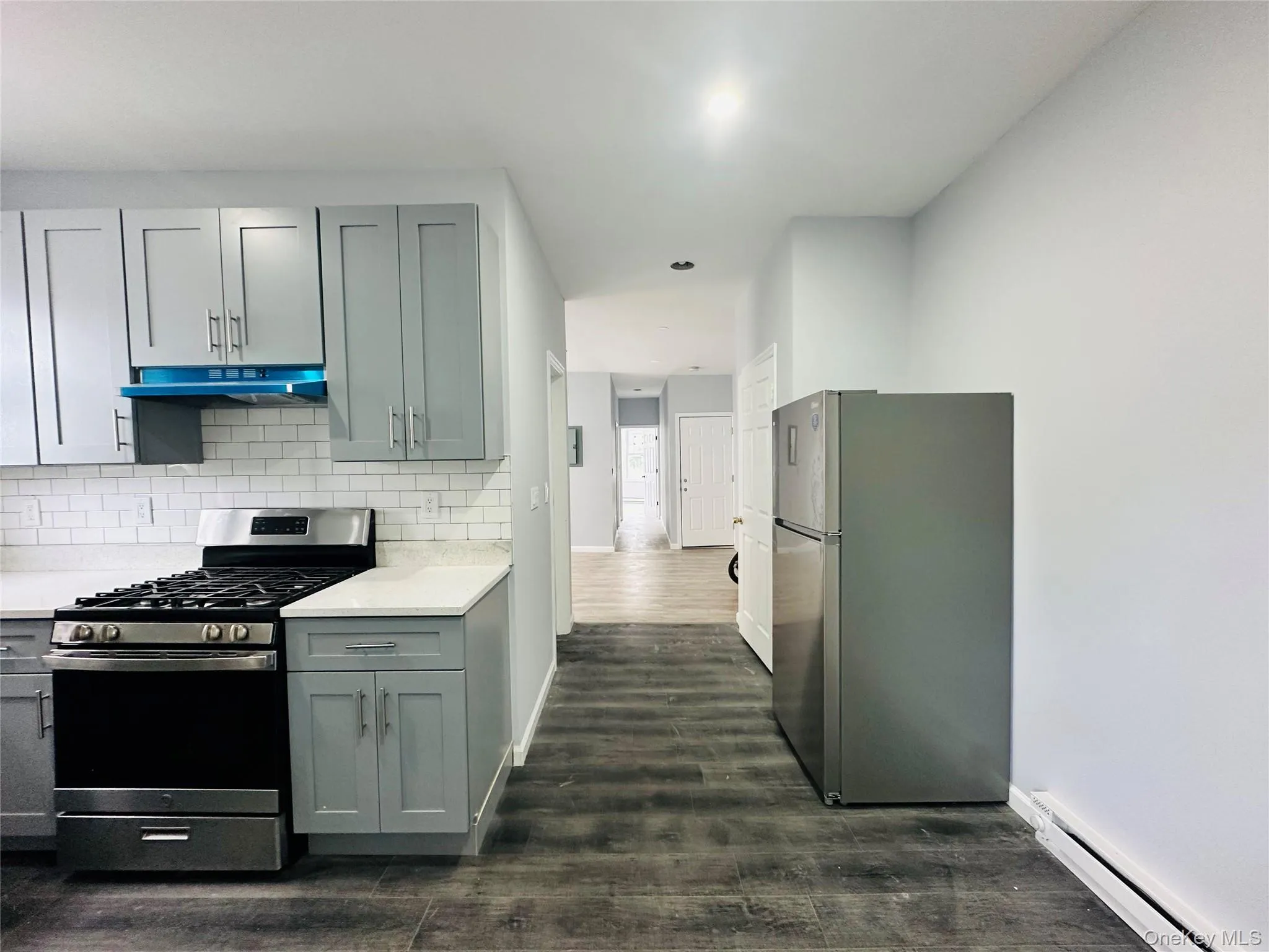 Kitchen featuring appliances with stainless steel finishes, baseboard heating, dark wood-style flooring, under cabinet range hood, and decorative backsplash Kitchen featuring appliances with stainless steel finishes, baseboard heating, dark wood-style flooring, under cabinet range hood, and decorative backsplash