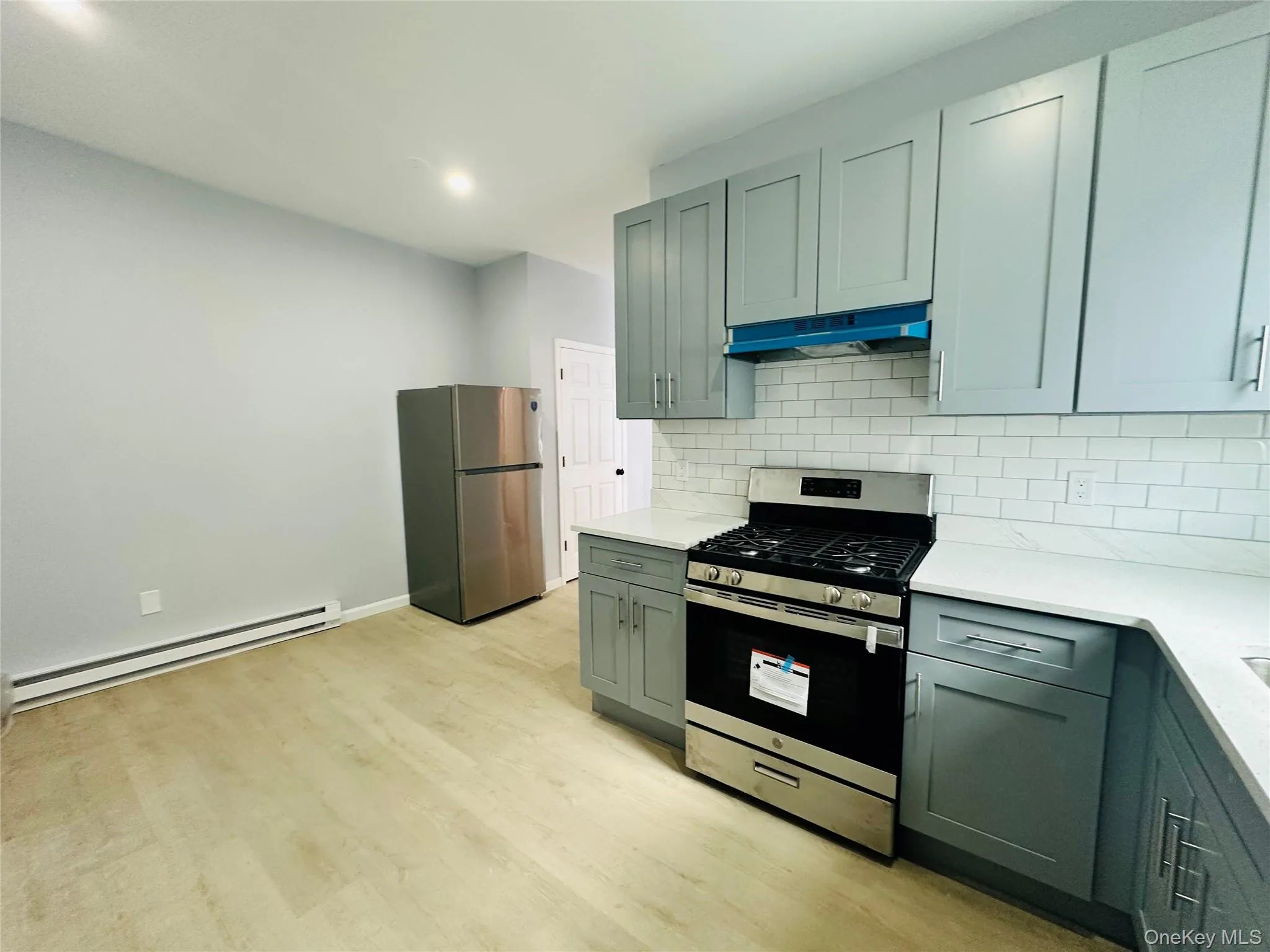 Kitchen featuring stainless steel appliances, a baseboard heating unit, backsplash, light wood-style flooring, and under cabinet range hood Kitchen featuring stainless steel appliances, a baseboard heating unit, backsplash, light wood-style flooring, and under cabinet range hood