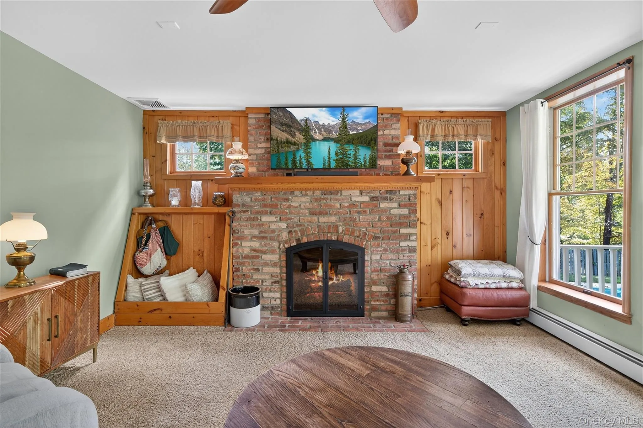 Carpeted living room featuring a brick fireplace, wooden walls, a baseboard radiator, and ceiling fan Carpeted living room featuring a brick fireplace, wooden walls, a baseboard radiator, and ceiling fan