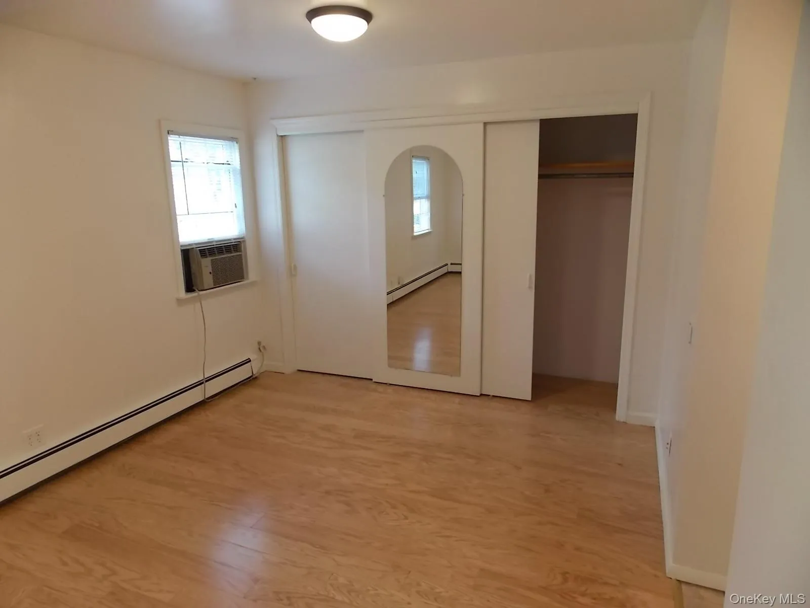 Unfurnished bedroom with arched walkways, light wood-style flooring, baseboard heating, two closets, and cooling unit Unfurnished bedroom with arched walkways, light wood-style flooring, baseboard heating, two closets, and cooling unit