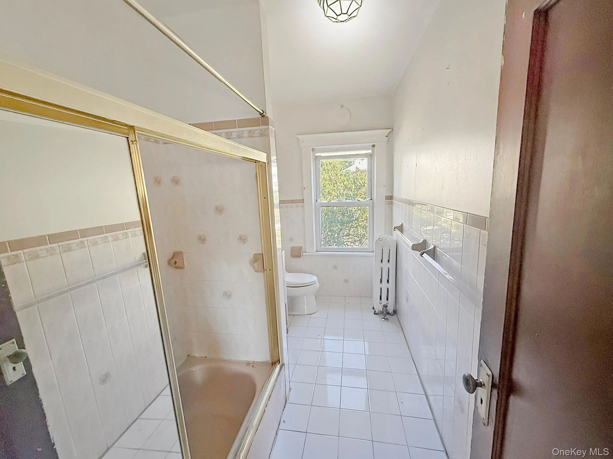 Bathroom with tile walls, bath / shower combo with glass door, light tile patterned flooring, a wainscoted wall, and radiator heating unit Bathroom with tile walls, bath / shower combo with glass door, light tile patterned flooring, a wainscoted wall, and radiator heating unit