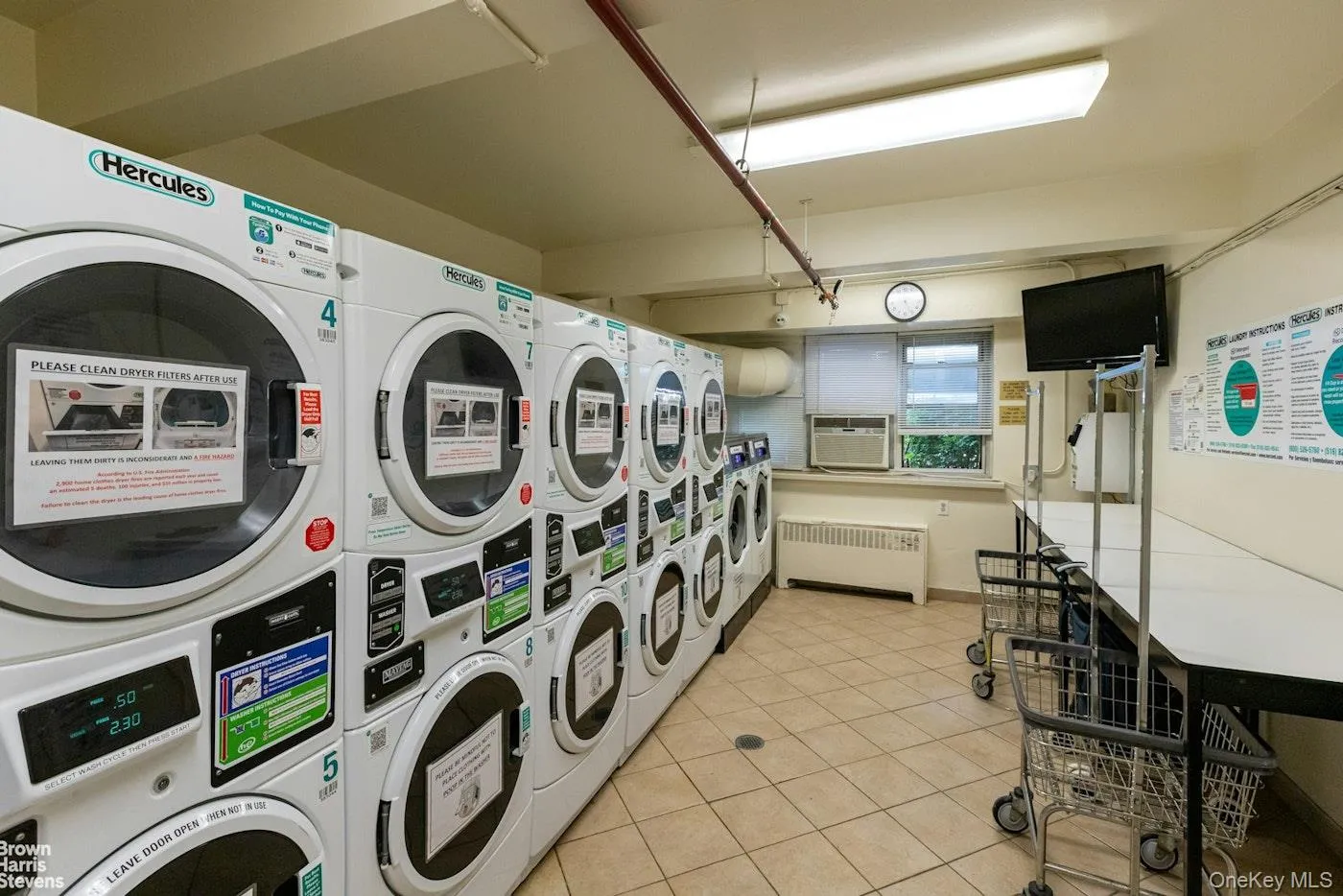 Community laundry room featuring radiator heating unit, light tile patterned flooring, stacked washer and clothes dryer, and cooling unit Community laundry room featuring radiator heating unit, light tile patterned flooring, stacked washer and clothes dryer, and cooling unit