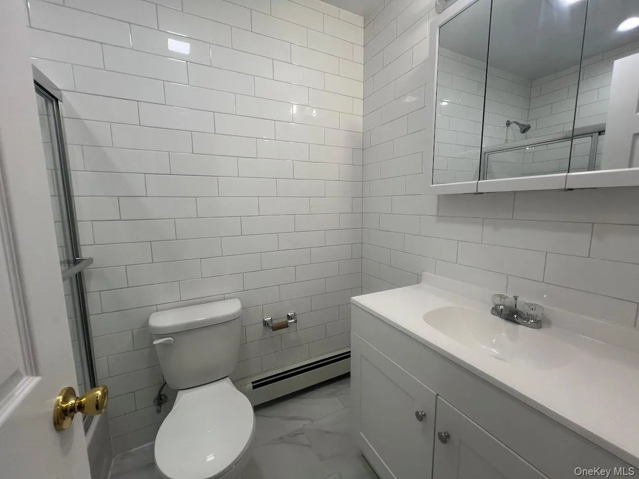 Full bathroom featuring vanity, tiled shower, tile walls, a baseboard radiator, and light marble finish floors Full bathroom featuring vanity, tiled shower, tile walls, a baseboard radiator, and light marble finish floors