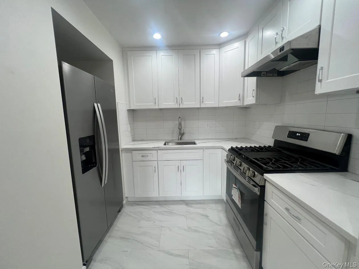 Kitchen featuring stainless steel appliances, tasteful backsplash, white cabinetry, under cabinet range hood, and light marble finish flooring Kitchen featuring stainless steel appliances, tasteful backsplash, white cabinetry, under cabinet range hood, and light marble finish flooring