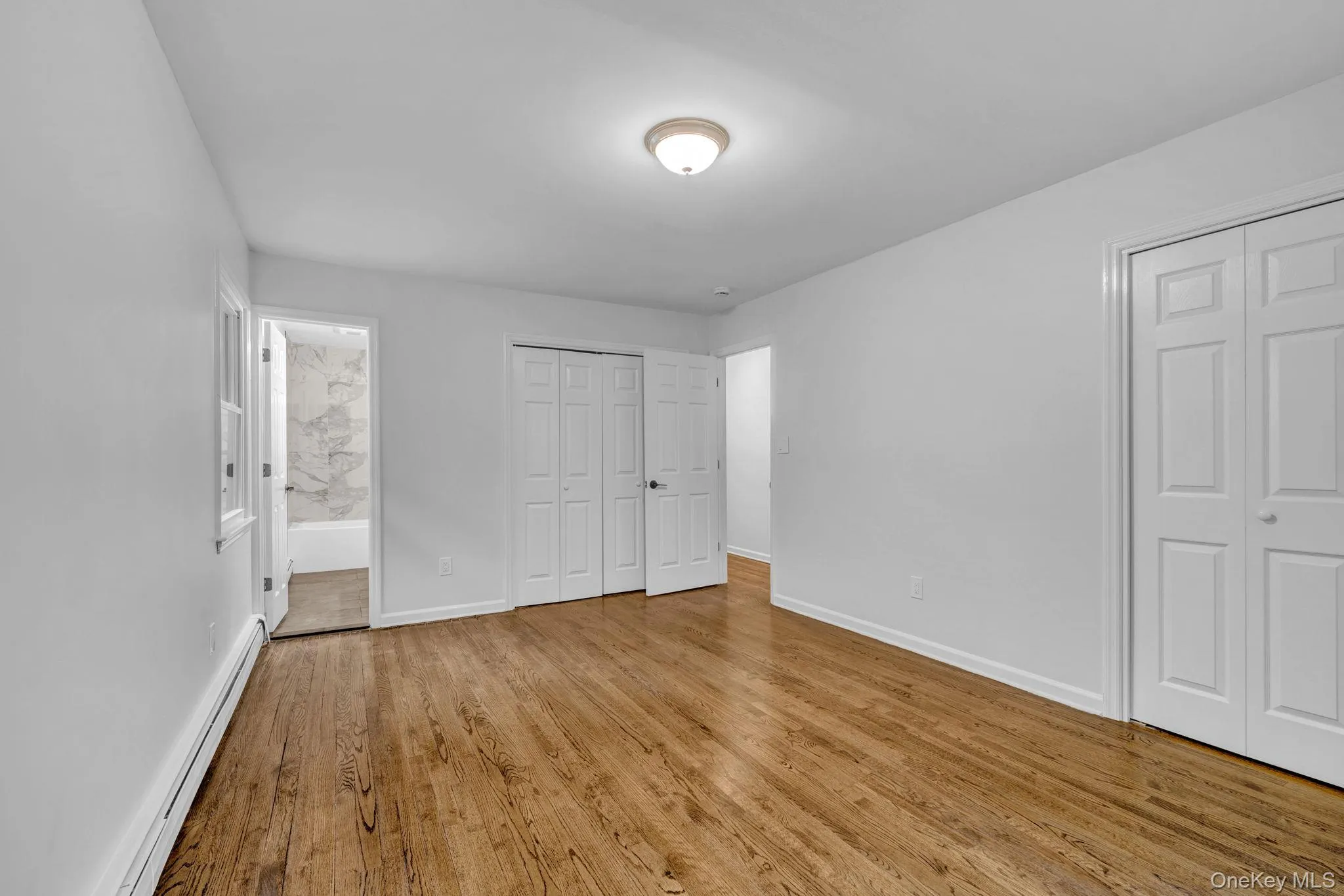 Unfurnished bedroom featuring multiple closets, a baseboard radiator, ensuite bath, and light wood-style floors Unfurnished bedroom featuring multiple closets, a baseboard radiator, ensuite bath, and light wood-style floors