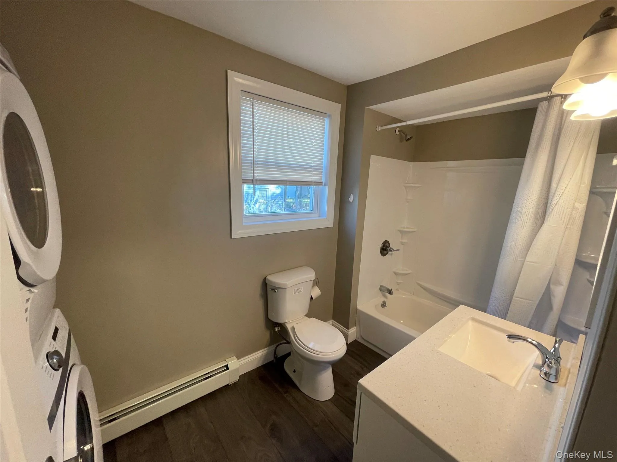 Bathroom featuring a baseboard heating unit, dark wood-style floors, shower / bath combo with shower curtain, vanity, and stacked washer / dryer Bathroom featuring a baseboard heating unit, dark wood-style floors, shower / bath combo with shower curtain, vanity, and stacked washer / dryer