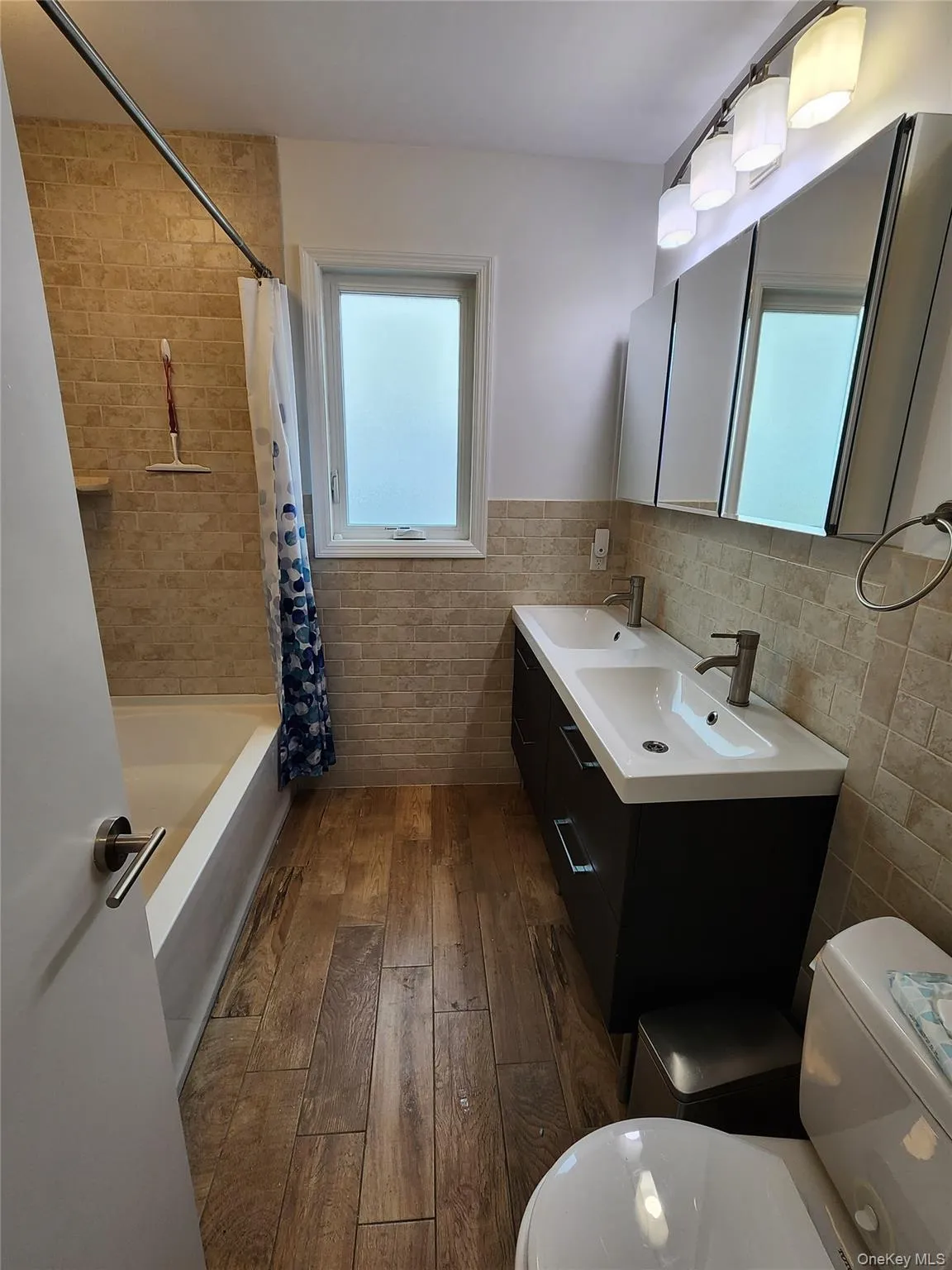 Bathroom featuring dark wood-style floors, tile walls, shower / bath combo with shower curtain, double vanity, and wainscoting Bathroom featuring dark wood-style floors, tile walls, shower / bath combo with shower curtain, double vanity, and wainscoting