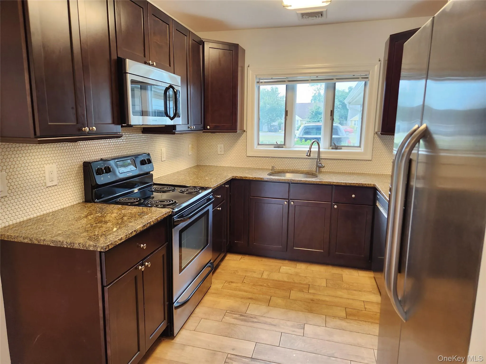 Kitchen with dark brown cabinetry, stainless steel appliances, light stone counters, and light wood-style flooring Kitchen with dark brown cabinetry, stainless steel appliances, light stone counters, and light wood-style flooring