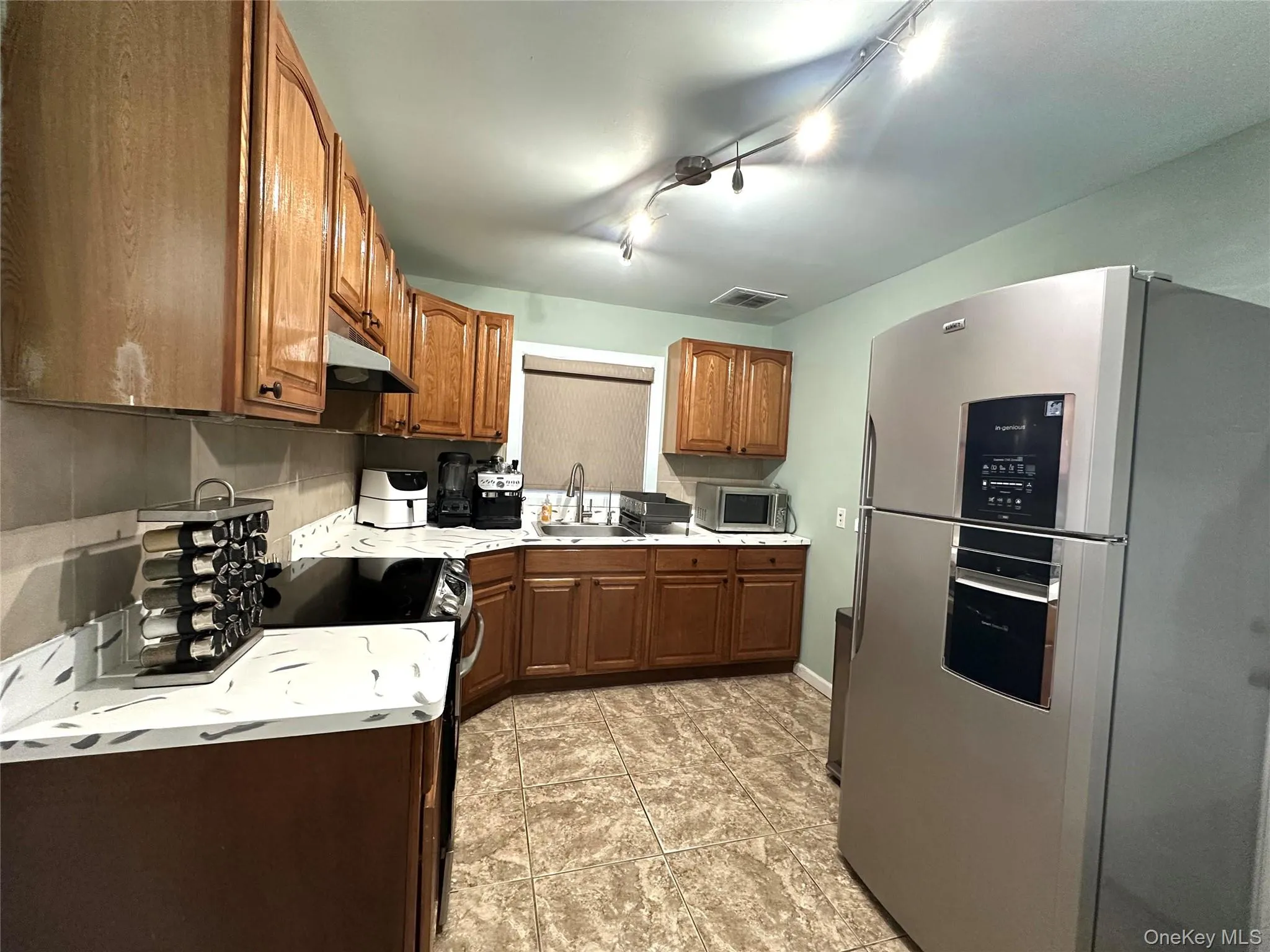 Kitchen with appliances with stainless steel finishes, brown cabinetry, track lighting, light countertops, and under cabinet range hood Kitchen with appliances with stainless steel finishes, brown cabinetry, track lighting, light countertops, and under cabinet range hood