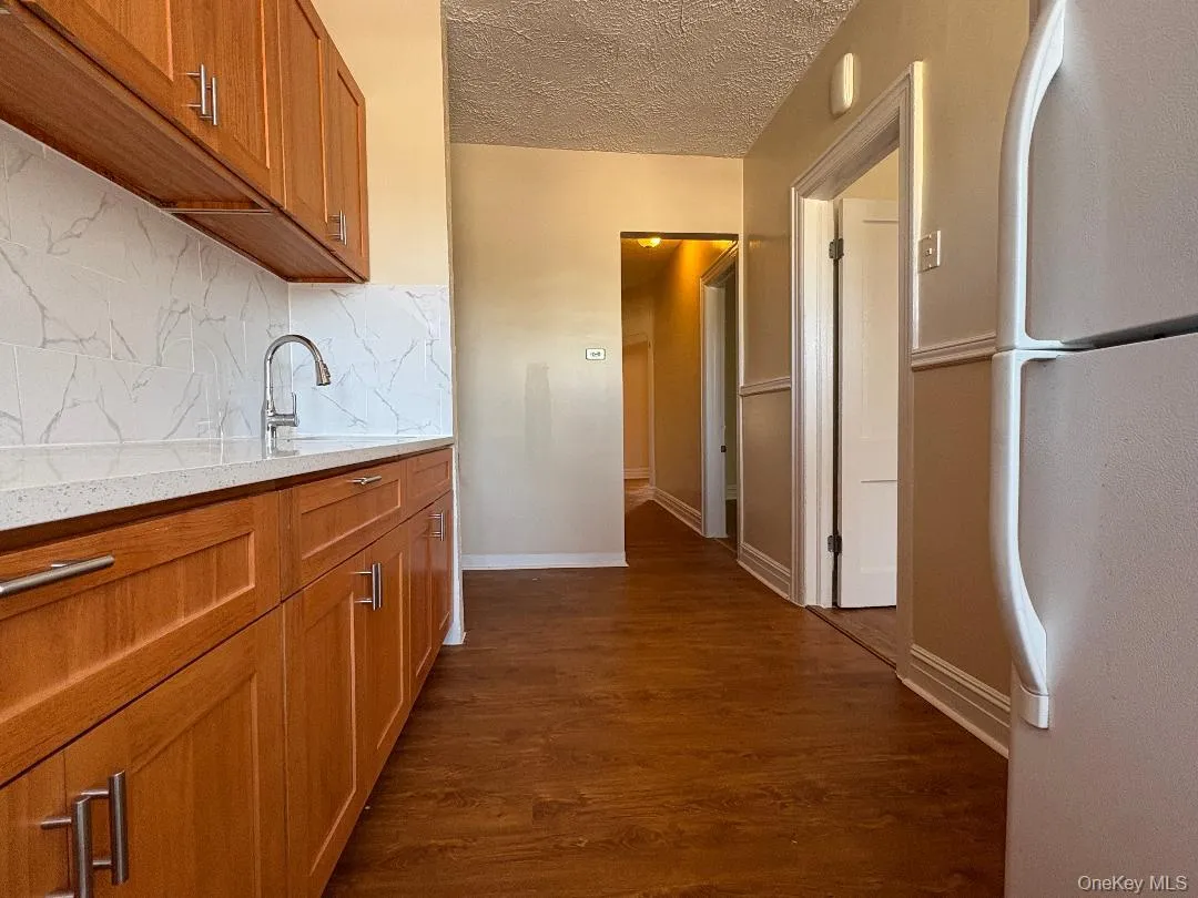 Kitchen featuring brown cabinets, freestanding refrigerator, decorative backsplash, dark wood-type flooring, and light stone counters Kitchen featuring brown cabinets, freestanding refrigerator, decorative backsplash, dark wood-type flooring, and light stone counters
