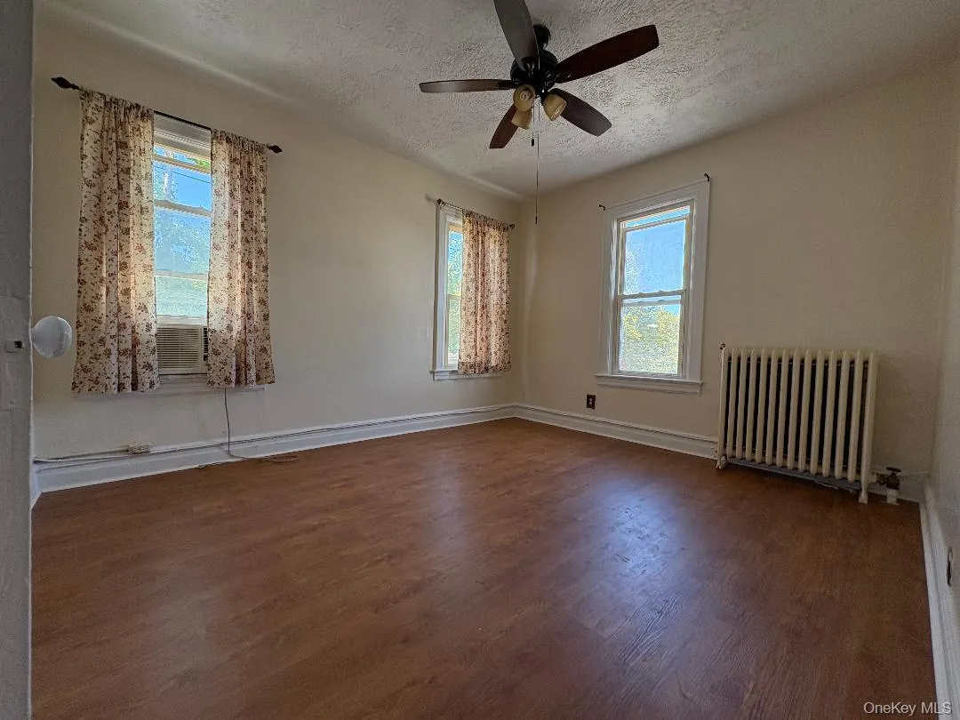 Empty room featuring radiator, a textured ceiling, wood finished floors, plenty of natural light, and ceiling fan Empty room featuring radiator, a textured ceiling, wood finished floors, plenty of natural light, and ceiling fan