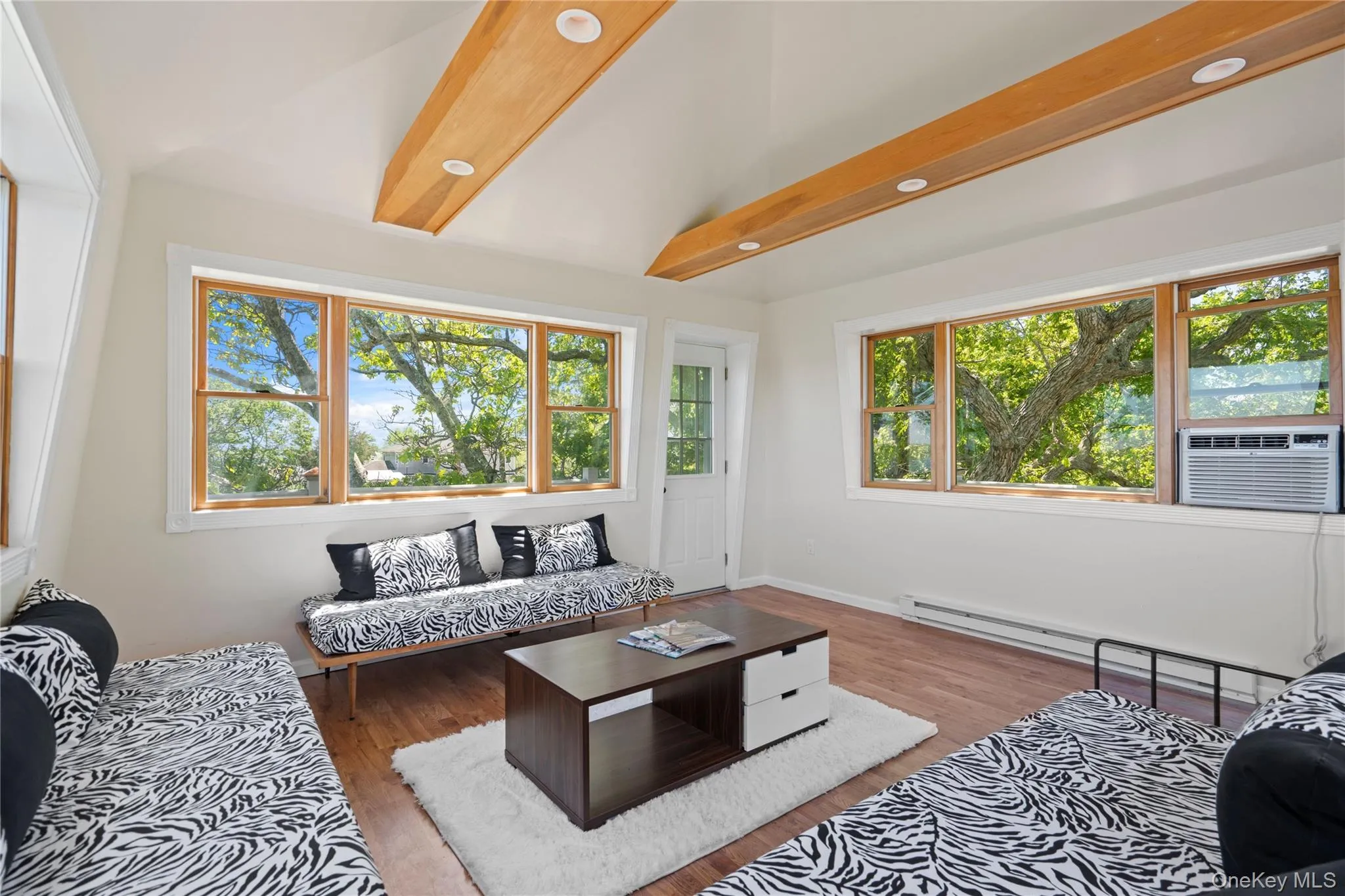 Living room featuring beam ceiling, plenty of natural light, wood finished floors, baseboard heating, and cooling unit Living room featuring beam ceiling, plenty of natural light, wood finished floors, baseboard heating, and cooling unit