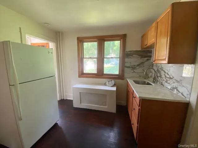 Kitchen featuring freestanding refrigerator, dark wood-style flooring, light stone countertops, tasteful backsplash, and brown cabinetry Kitchen featuring freestanding refrigerator, dark wood-style flooring, light stone countertops, tasteful backsplash, and brown cabinetry