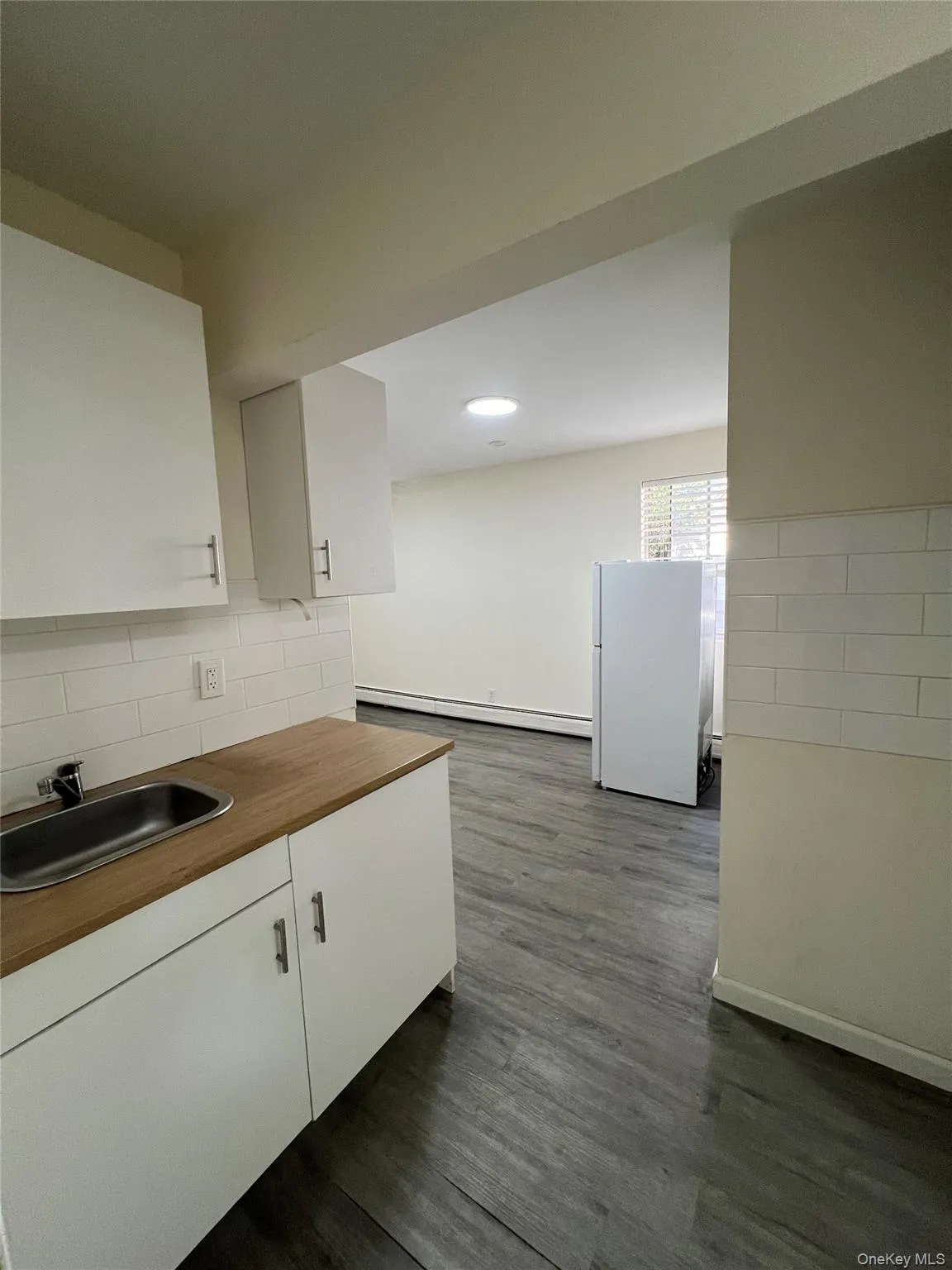 White cabinets, dark wood-type flooring, freestanding refrigerator, a baseboard heating unit, and decorative backsplash White cabinets, dark wood-type flooring, freestanding refrigerator, a baseboard heating unit, and decorative backsplash