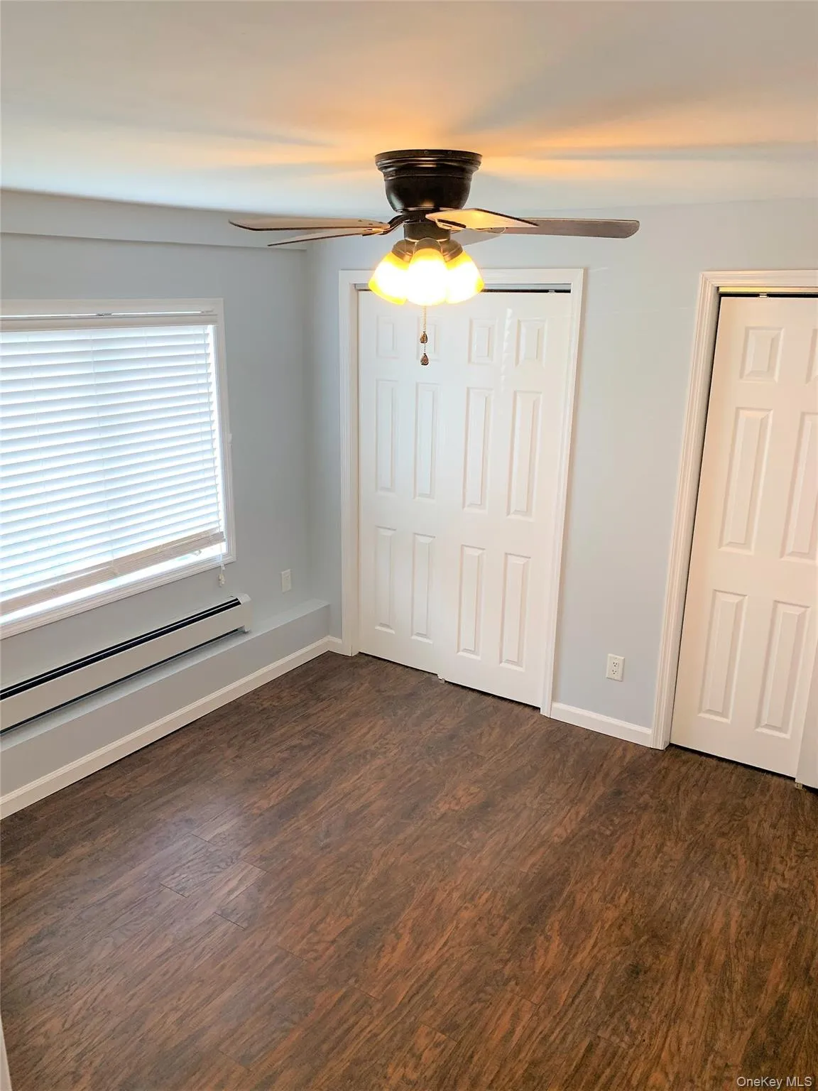 Unfurnished bedroom with a baseboard radiator, ceiling fan, dark wood-style floors, and multiple closets Unfurnished bedroom with a baseboard radiator, ceiling fan, dark wood-style floors, and multiple closets