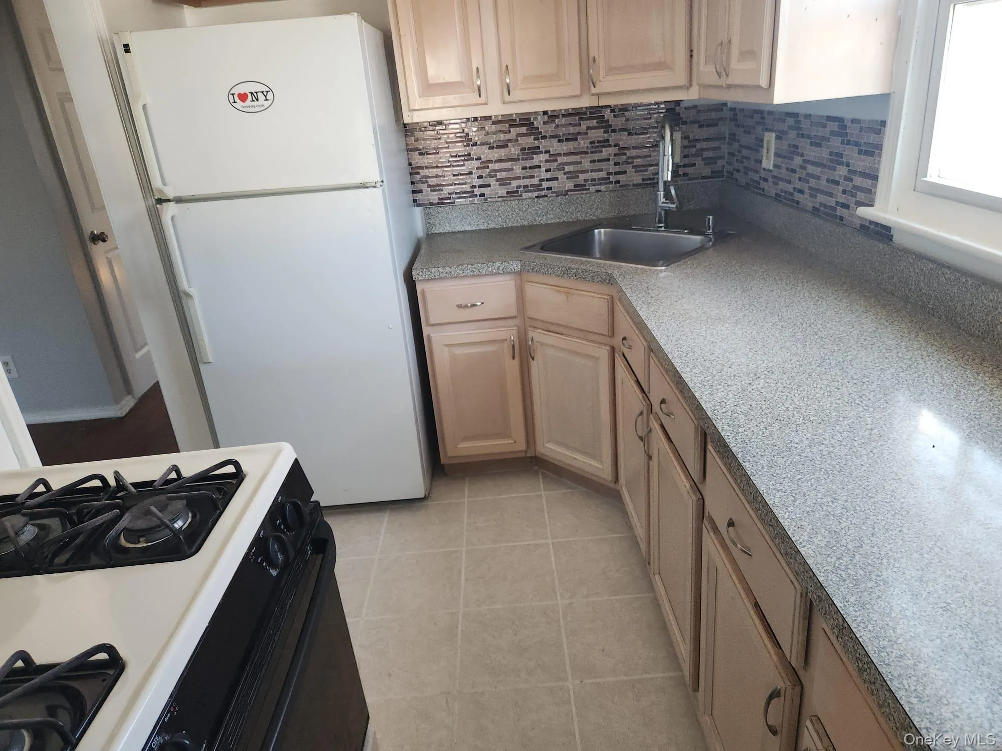 Kitchen with freestanding refrigerator, black gas range oven, backsplash, light tile patterned floors, and light brown cabinets Kitchen with freestanding refrigerator, black gas range oven, backsplash, light tile patterned floors, and light brown cabinets