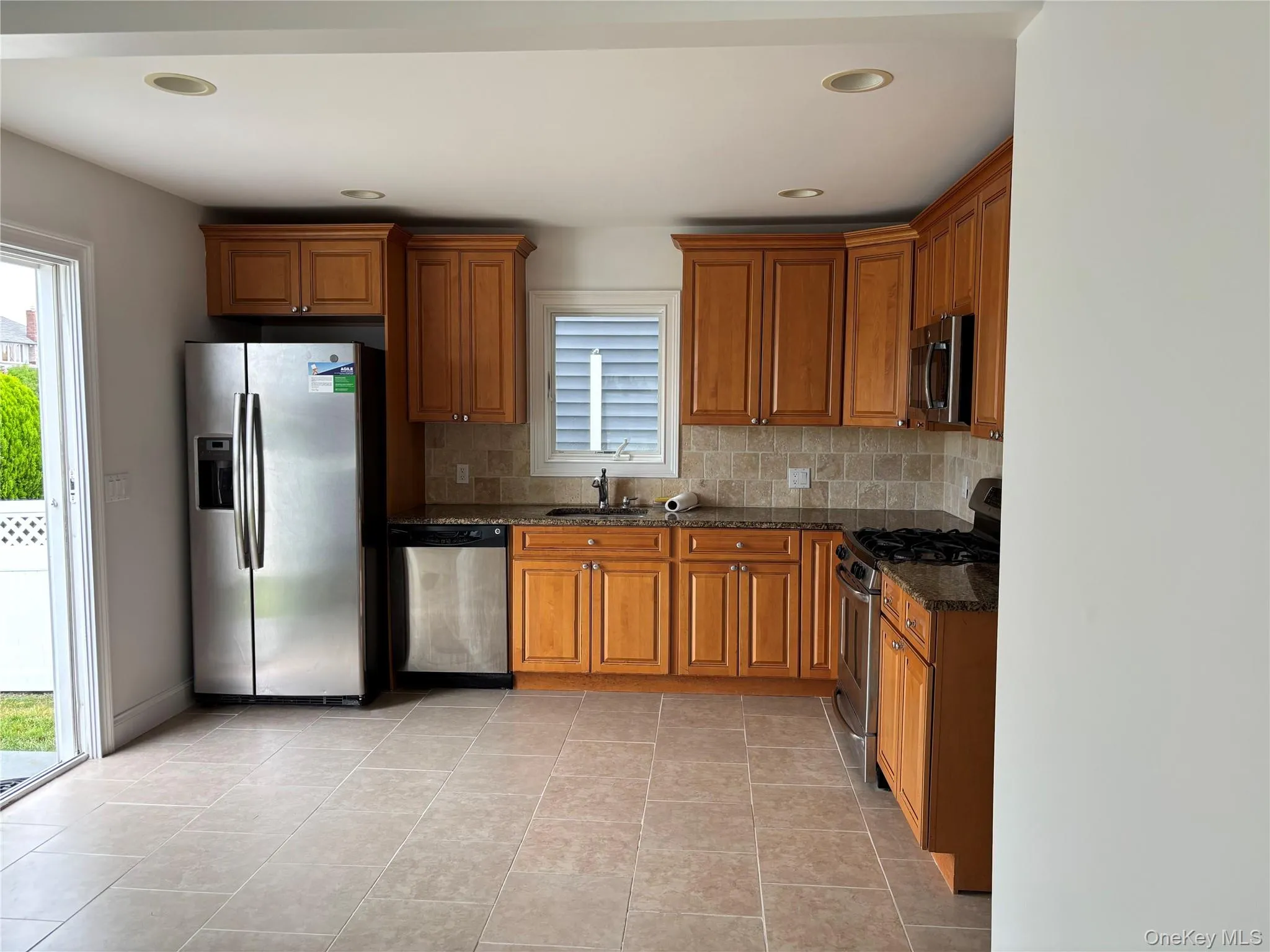 Kitchen with stainless steel appliances, brown cabinets, decorative backsplash, light tile patterned floors, and dark stone counters Kitchen with stainless steel appliances, brown cabinets, decorative backsplash, light tile patterned floors, and dark stone counters