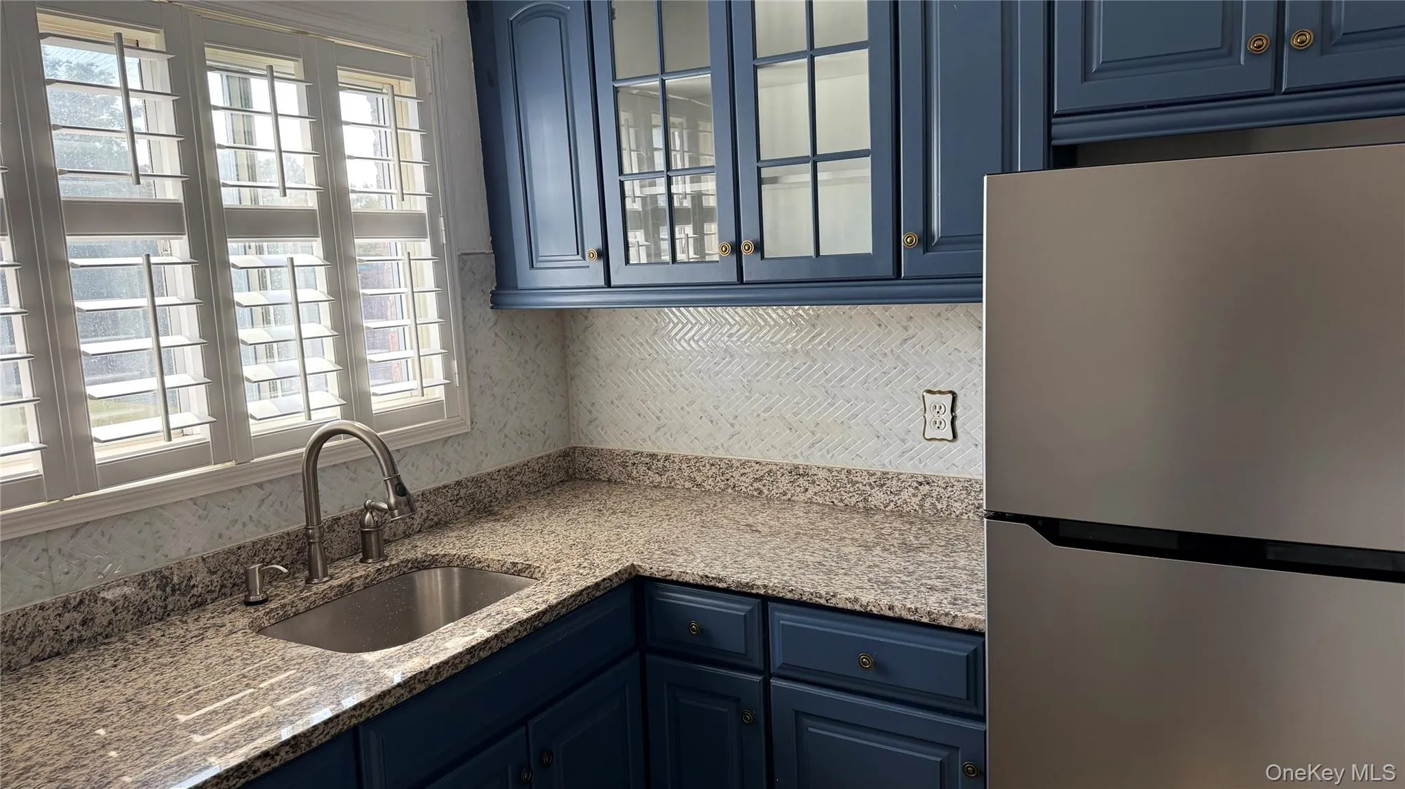 Kitchen with blue cabinets, freestanding refrigerator, light stone countertops, and glass insert cabinets Kitchen with blue cabinets, freestanding refrigerator, light stone countertops, and glass insert cabinets