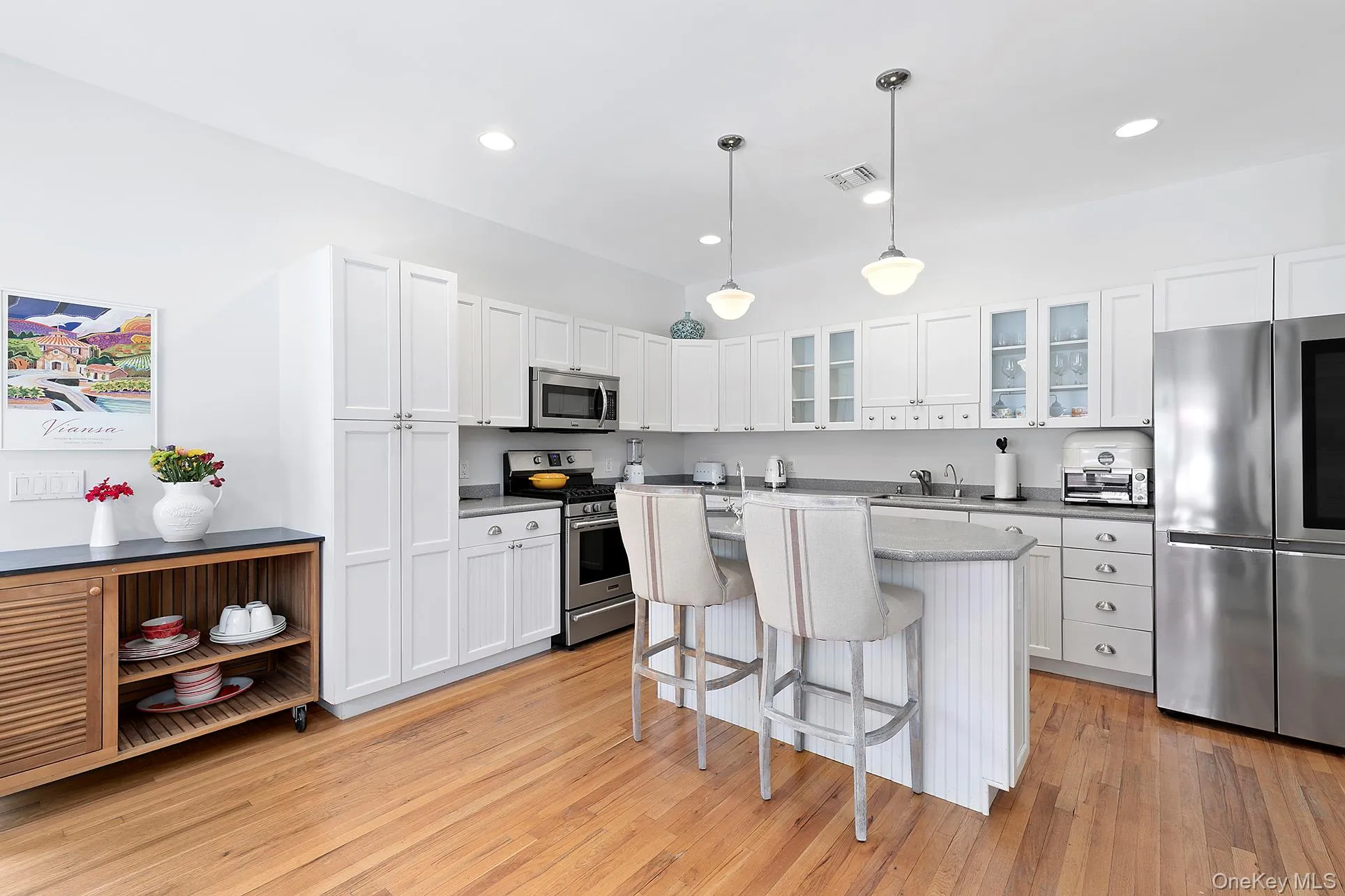 Kitchen featuring white cabinetry, a breakfast bar, appliances with stainless steel finishes, decorative light fixtures, and glass insert cabinets Kitchen featuring white cabinetry, a breakfast bar, appliances with stainless steel finishes, decorative light fixtures, and glass insert cabinets