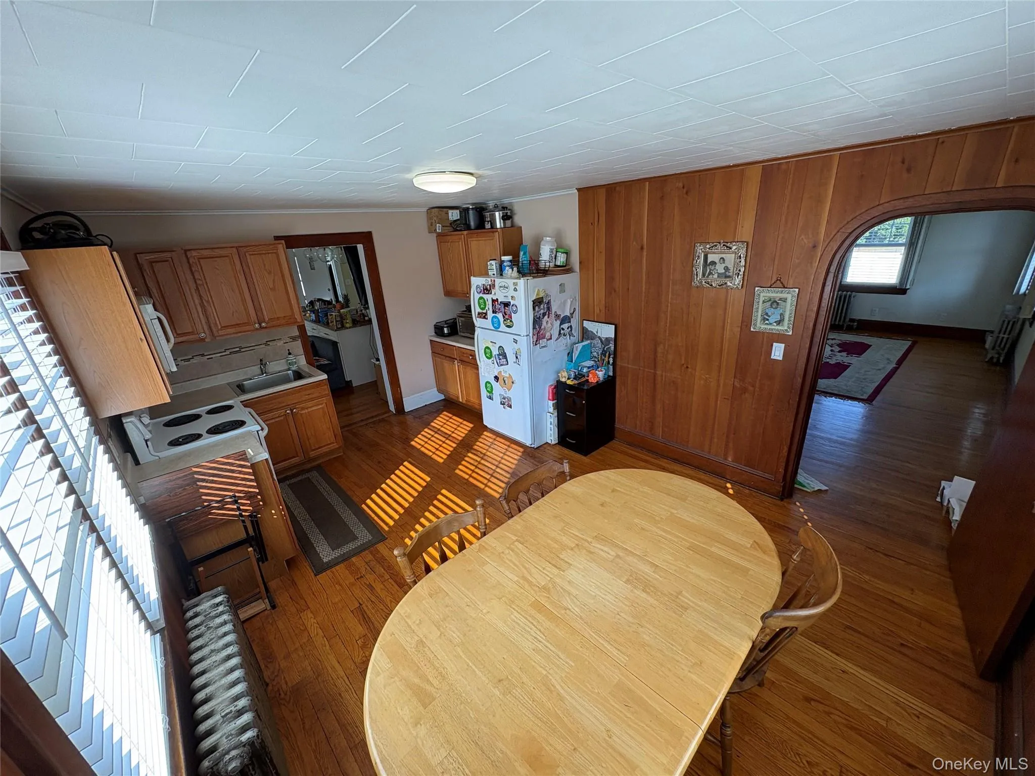 Kitchen featuring arched walkways, wood walls, light wood-type flooring, freestanding refrigerator, and brown cabinetry Kitchen featuring arched walkways, wood walls, light wood-type flooring, freestanding refrigerator, and brown cabinetry