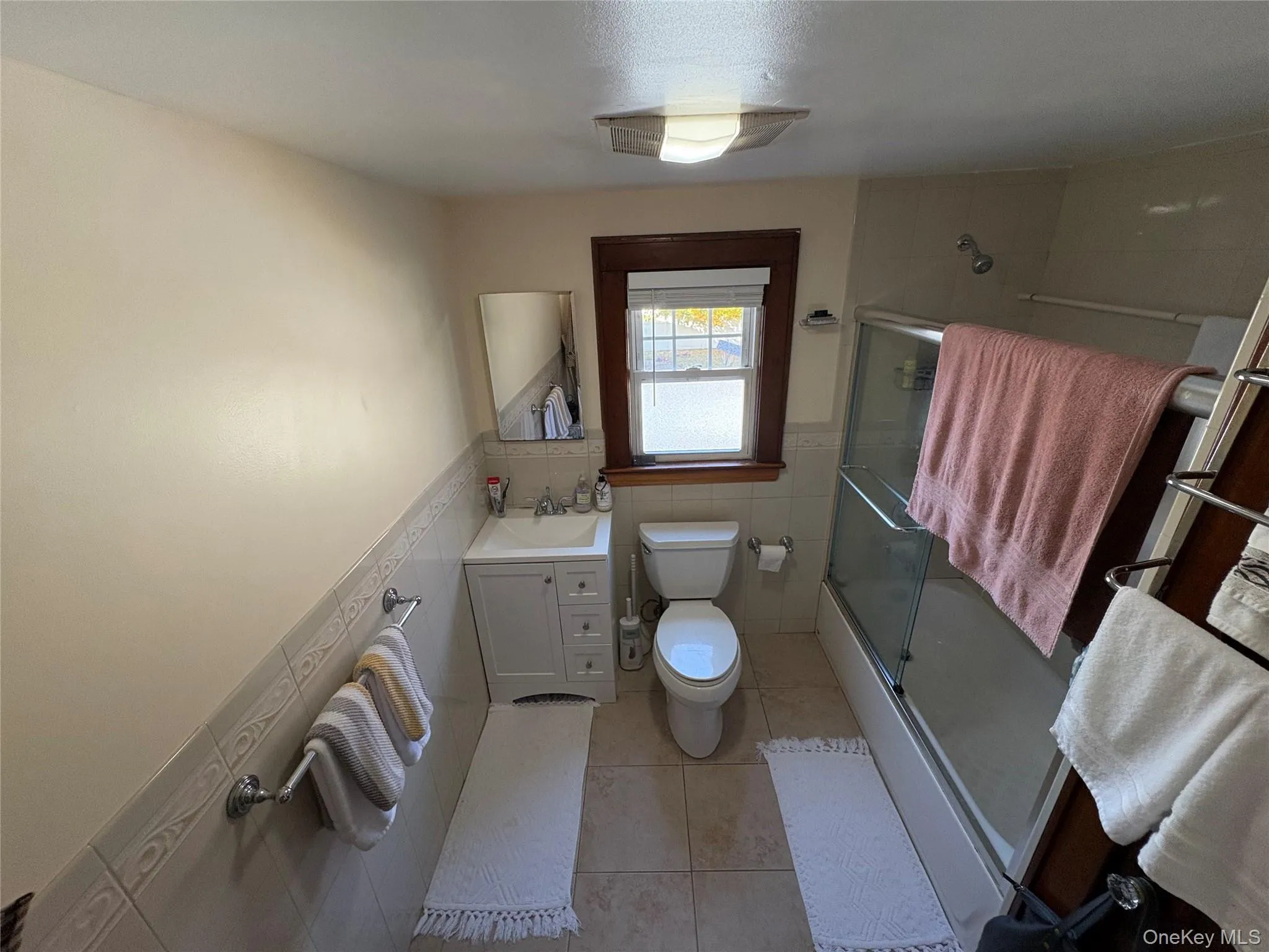Full bathroom with vanity, tile walls, light tile patterned floors, shower / bath combination with glass door, and a wainscoted wall Full bathroom with vanity, tile walls, light tile patterned floors, shower / bath combination with glass door, and a wainscoted wall