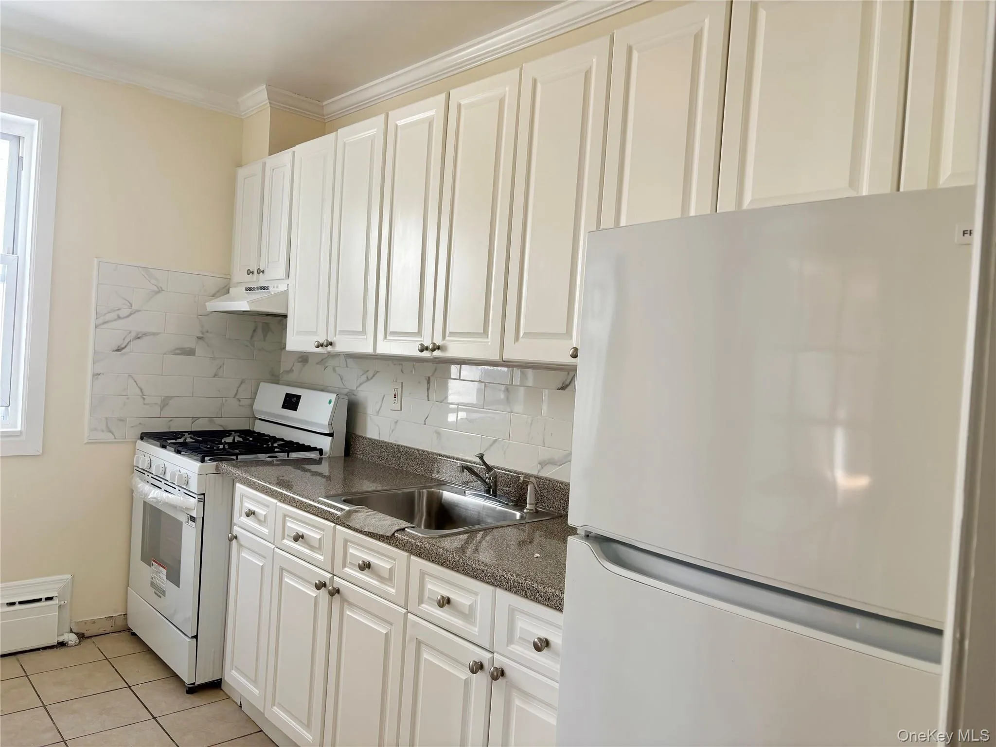 Kitchen with white appliances, decorative backsplash, light tile patterned floors, under cabinet range hood, and ornamental molding Kitchen with white appliances, decorative backsplash, light tile patterned floors, under cabinet range hood, and ornamental molding