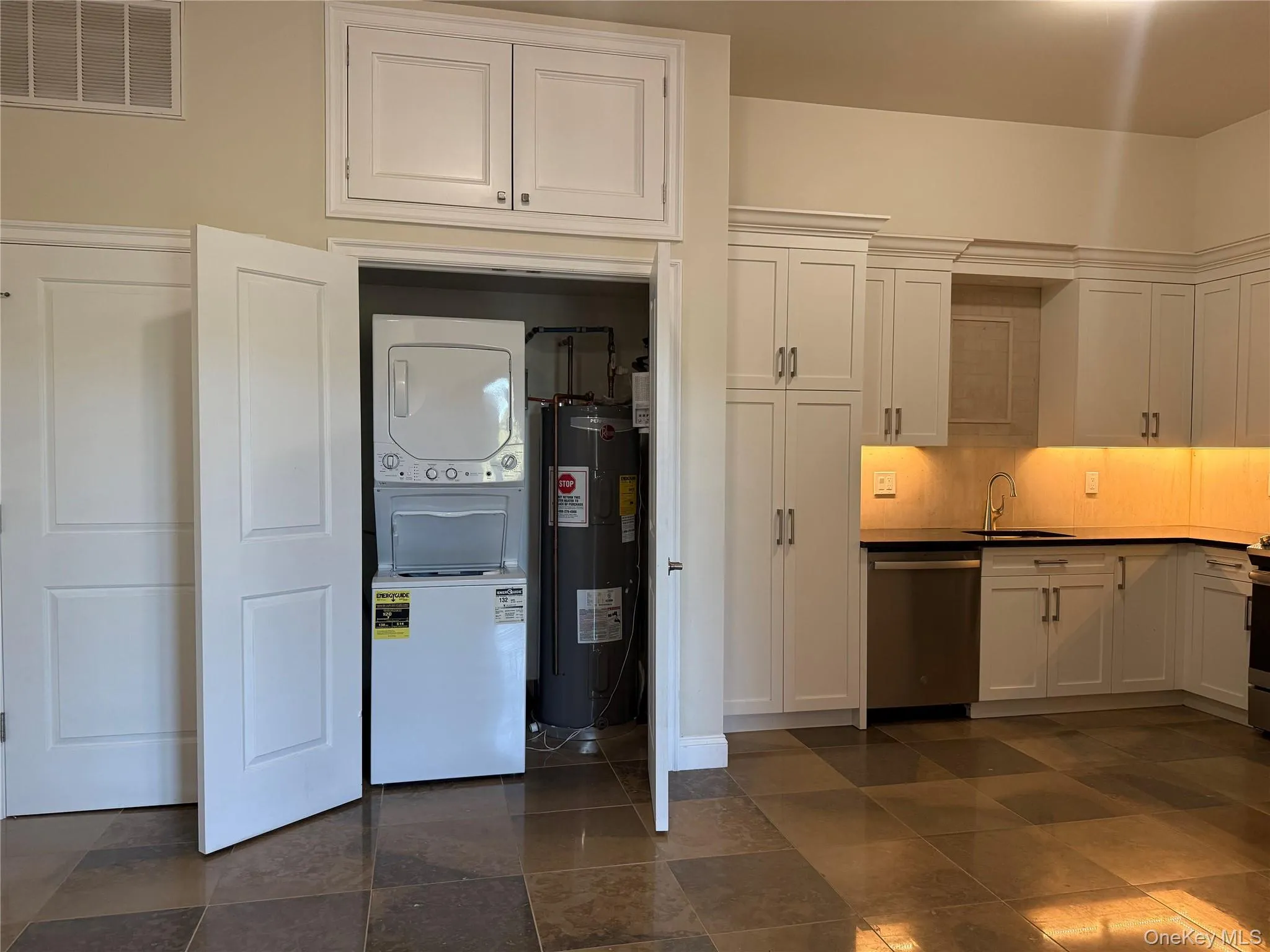 Kitchen featuring white cabinets, estacked washer and dryer, dark countertops, dishwasher, and water heater Kitchen featuring white cabinets, estacked washer and dryer, dark countertops, dishwasher, and water heater