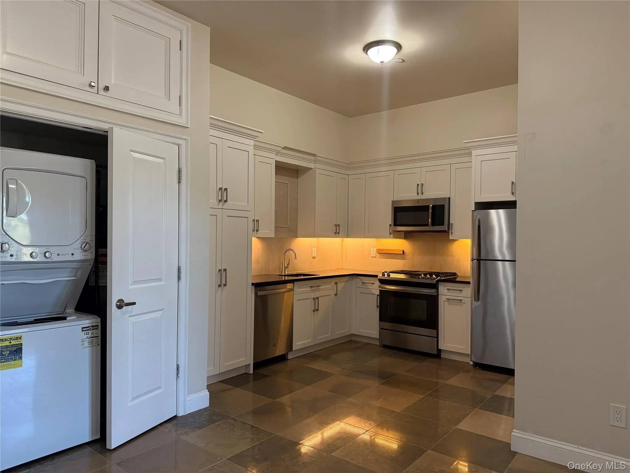Kitchen featuring white cabinetry, appliances with stainless steel finishes, dark countertops, backsplash, and stacked washer / dryer Kitchen featuring white cabinetry, appliances with stainless steel finishes, dark countertops, backsplash, and stacked washer / dryer