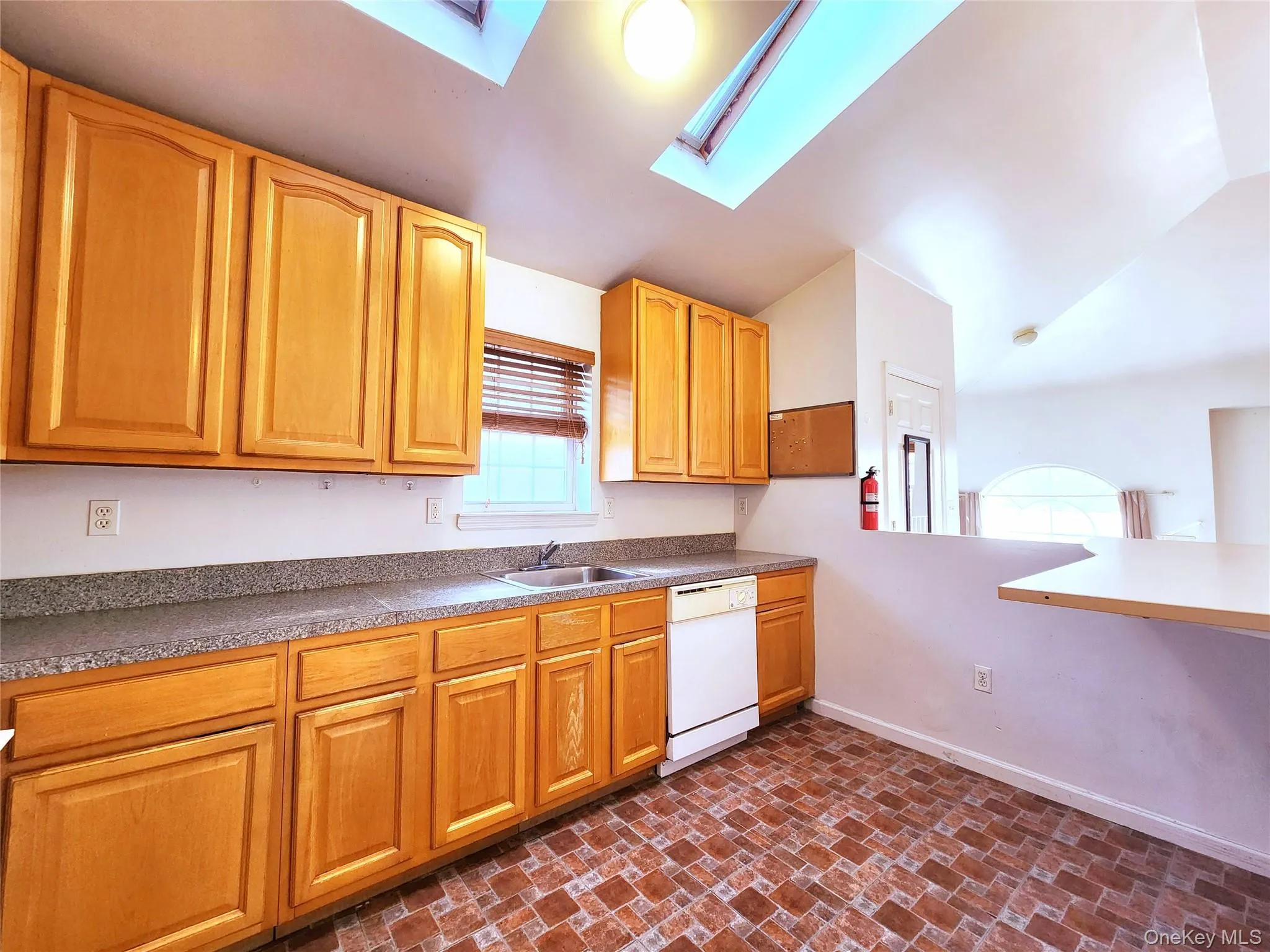 Kitchen featuring white dishwasher, brick patterned flooring, lofted ceiling, a skylight, and dark countertops Kitchen featuring white dishwasher, brick patterned flooring, lofted ceiling, a skylight, and dark countertops