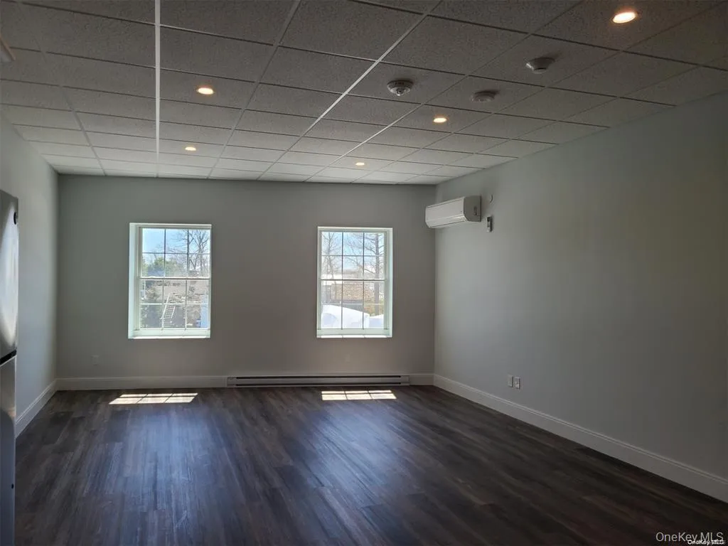 Empty room with a drop ceiling, dark wood-style flooring, baseboard heating, a wall unit AC, and recessed lighting Empty room with a drop ceiling, dark wood-style flooring, baseboard heating, a wall unit AC, and recessed lighting