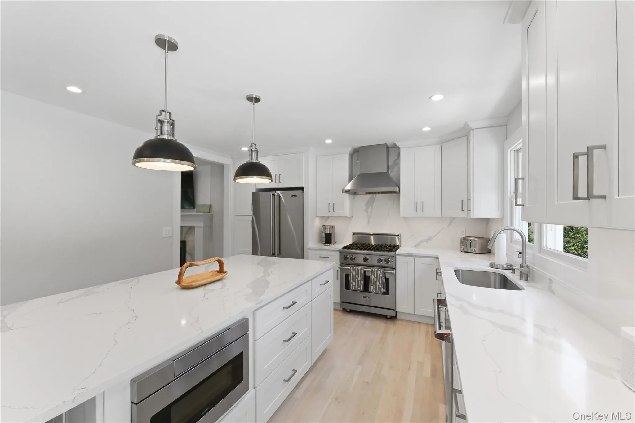 Kitchen with high end appliances, decorative light fixtures, wall chimney range hood, light wood-style flooring, and light stone counters Kitchen with high end appliances, decorative light fixtures, wall chimney range hood, light wood-style flooring, and light stone counters