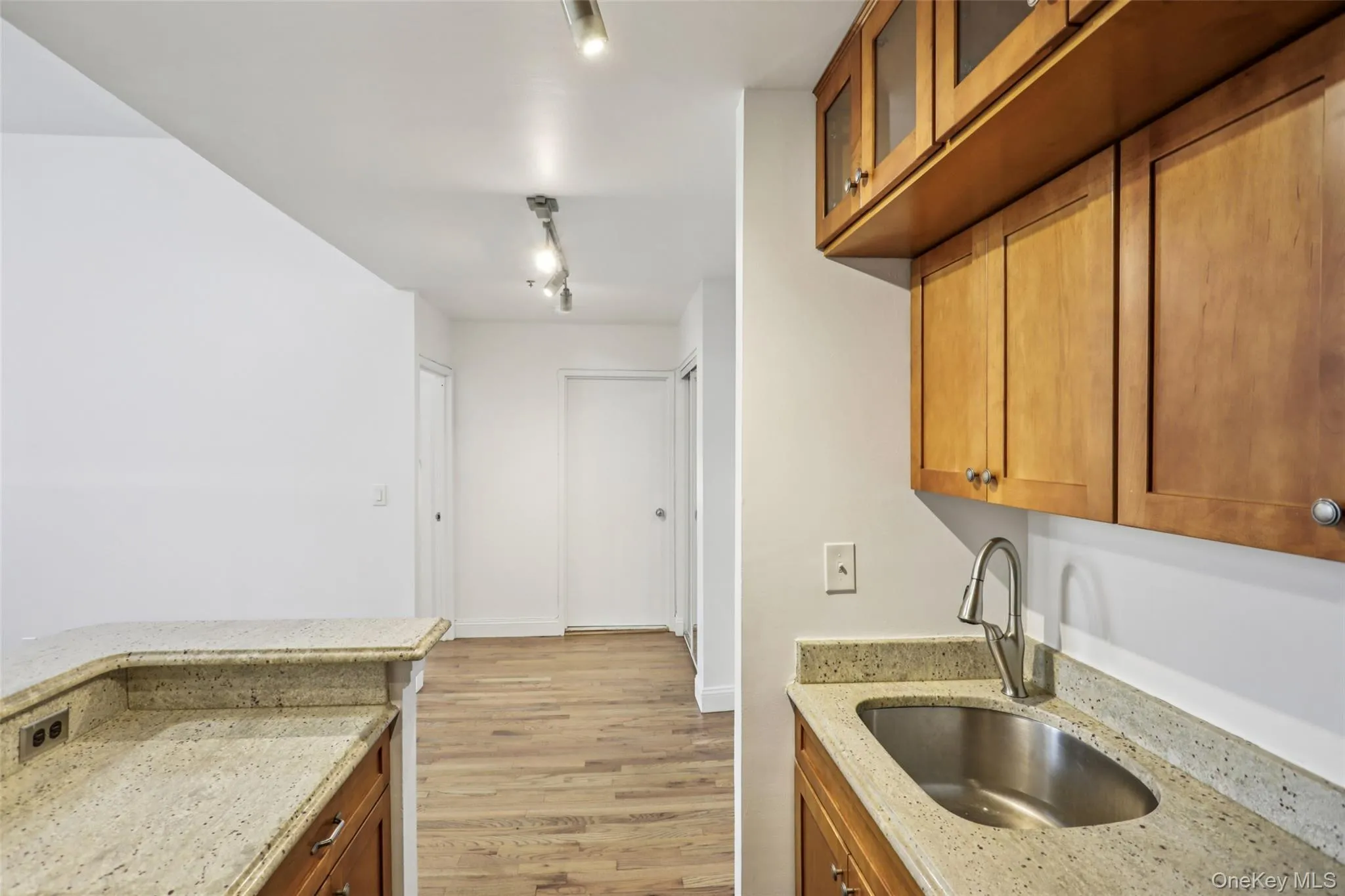Kitchen featuring brown cabinets, light stone countertops, light wood-type flooring, glass insert cabinets, and rail lighting Kitchen featuring brown cabinets, light stone countertops, light wood-type flooring, glass insert cabinets, and rail lighting
