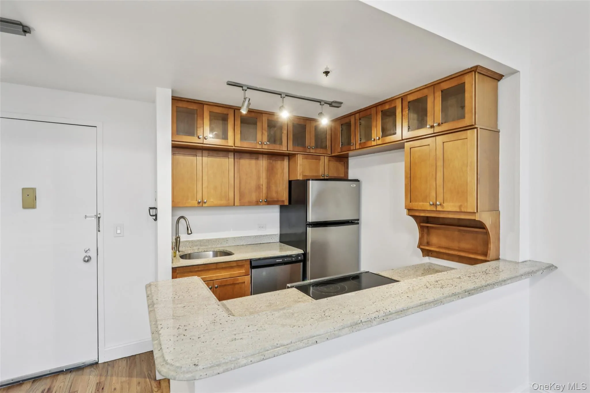 Kitchen with brown cabinets, light stone counters, glass insert cabinets, rail lighting, and appliances with stainless steel finishes Kitchen with brown cabinets, light stone counters, glass insert cabinets, rail lighting, and appliances with stainless steel finishes