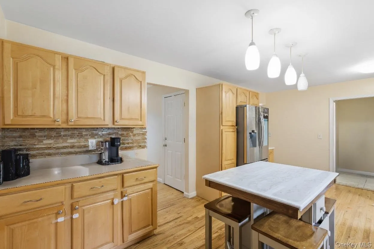 Kitchen with light countertops, decorative backsplash, stainless steel refrigerator with ice dispenser, decorative light fixtures, and light brown cabinets Kitchen with light countertops, decorative backsplash, stainless steel refrigerator with ice dispenser, decorative light fixtures, and light brown cabinets