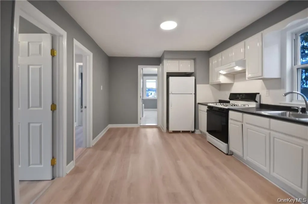 Kitchen featuring white cabinets, white appliances, dark countertops, light wood-type flooring, and under cabinet range hood Kitchen featuring white cabinets, white appliances, dark countertops, light wood-type flooring, and under cabinet range hood