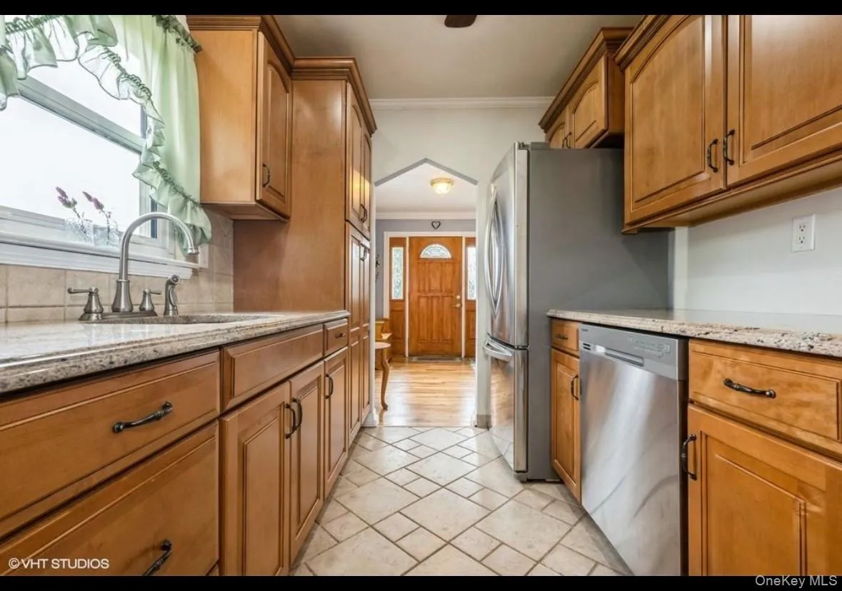 Kitchen with brown cabinetry, crown molding, light stone countertops, stainless steel dishwasher, and stone tile flooring Kitchen with brown cabinetry, crown molding, light stone countertops, stainless steel dishwasher, and stone tile flooring