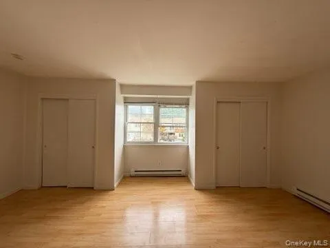 Unfurnished bedroom with two closets, light wood-type flooring, and a baseboard radiator Unfurnished bedroom with two closets, light wood-type flooring, and a baseboard radiator