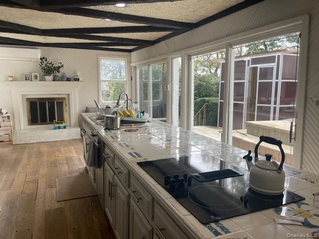 Kitchen with tile counters, black electric stovetop, beamed ceiling, dark wood finished floors, and a brick fireplace Kitchen with tile counters, black electric stovetop, beamed ceiling, dark wood finished floors, and a brick fireplace