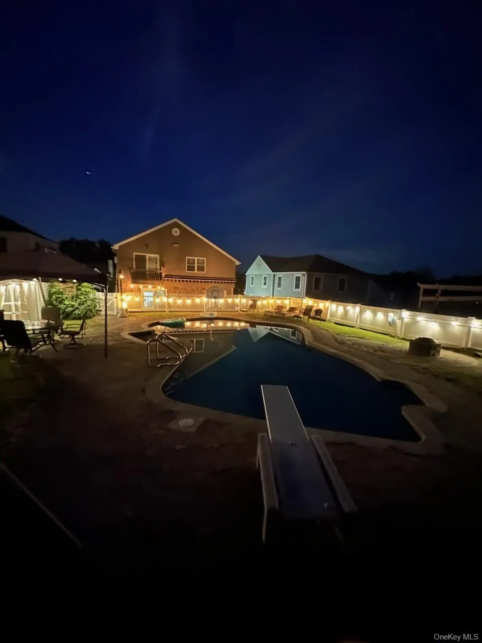 Pool at night with a patio area, a diving board, showing fire pit Pool at night with a patio area, a diving board, showing fire pit