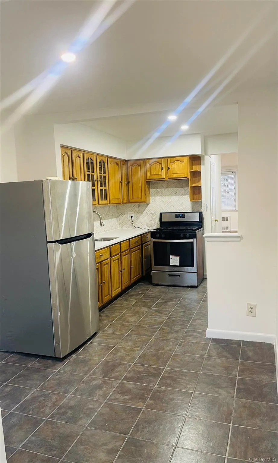 Kitchen featuring appliances with stainless steel finishes, tasteful backsplash, light countertops, brown cabinets, and glass insert cabinets Kitchen featuring appliances with stainless steel finishes, tasteful backsplash, light countertops, brown cabinets, and glass insert cabinets