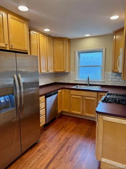 Kitchen featuring dark countertops, stainless steel appliances, dark wood-style flooring, decorative backsplash, and light brown cabinetry Kitchen featuring dark countertops, stainless steel appliances, dark wood-style flooring, decorative backsplash, and light brown cabinetry