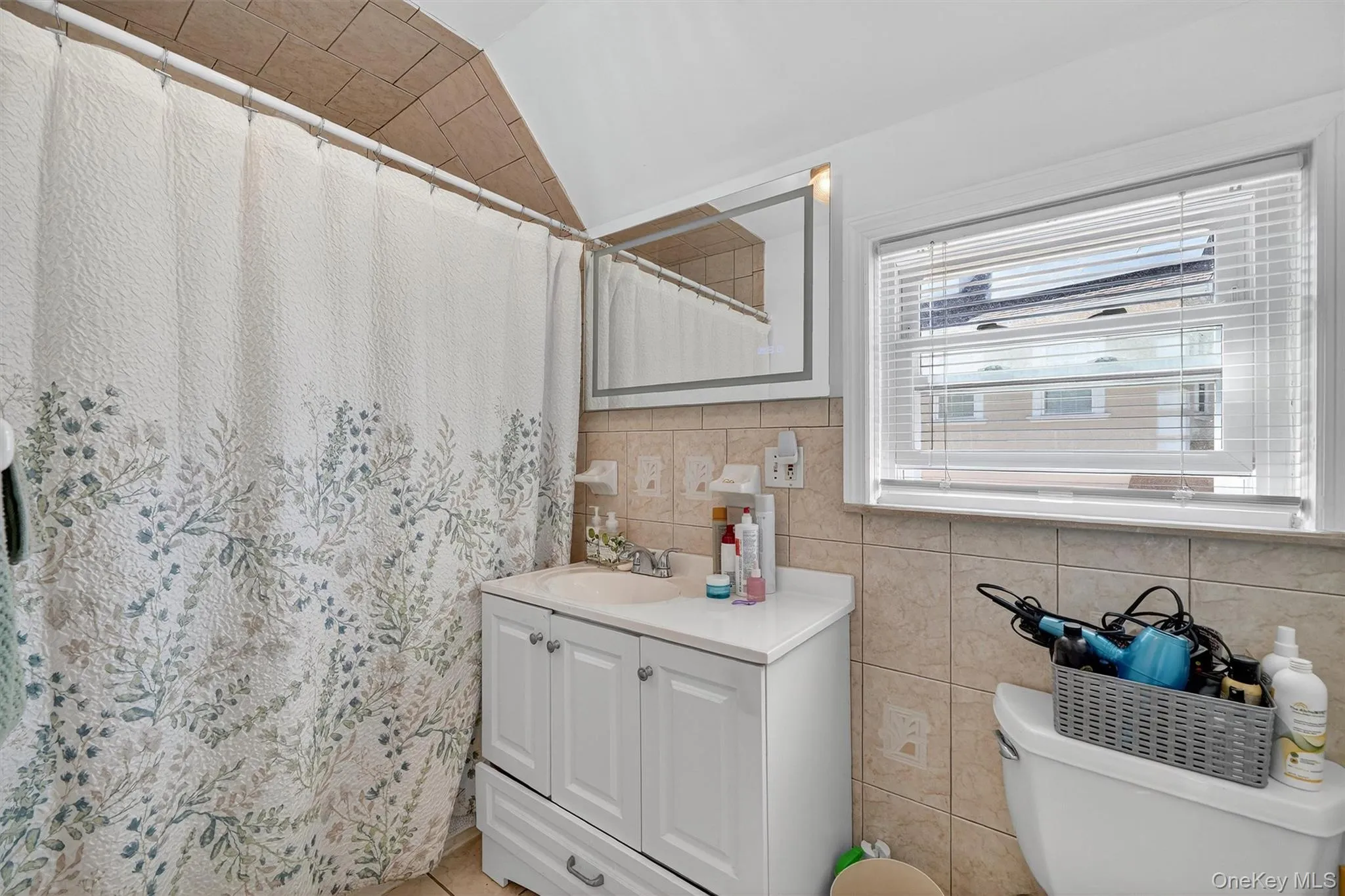 Bathroom with vanity, tile walls, curtained shower, vaulted ceiling, and decorative backsplash Bathroom with vanity, tile walls, curtained shower, vaulted ceiling, and decorative backsplash
