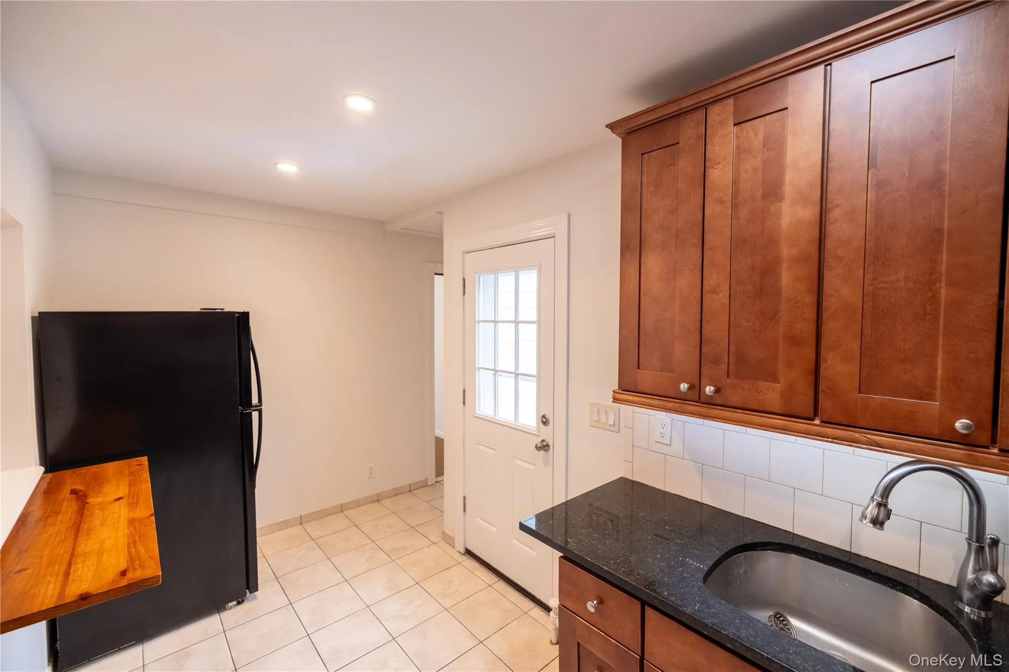 Kitchen featuring dark stone countertops, freestanding refrigerator, brown cabinetry, decorative backsplash, and light tile patterned floors Kitchen featuring dark stone countertops, freestanding refrigerator, brown cabinetry, decorative backsplash, and light tile patterned floors