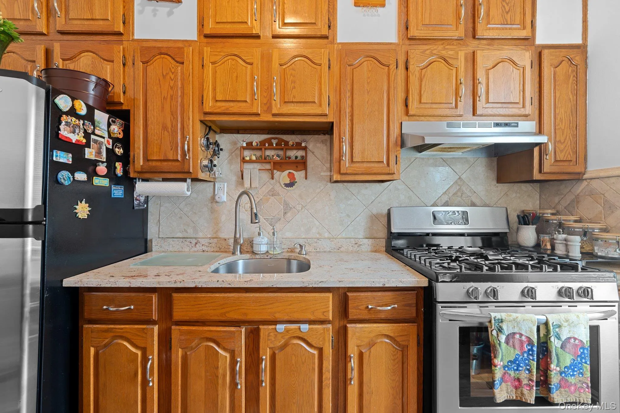 Kitchen with stainless steel appliances, backsplash, light stone counters, brown cabinetry, and under cabinet range hood Kitchen with stainless steel appliances, backsplash, light stone counters, brown cabinetry, and under cabinet range hood