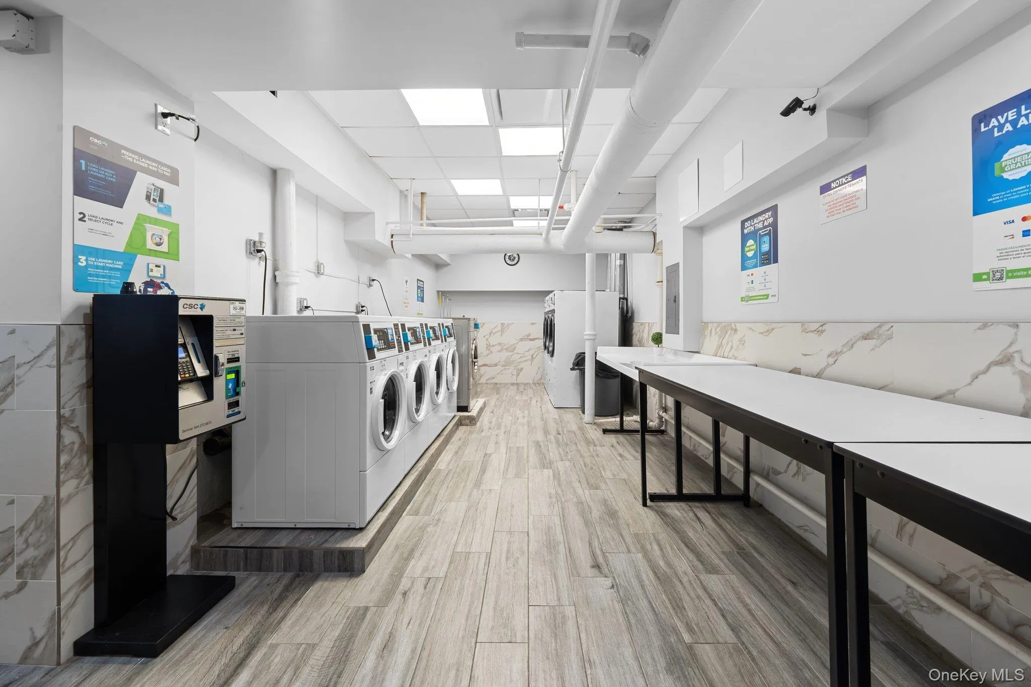 Community laundry room with tile walls, independent washer and dryer, light wood-style flooring, a wainscoted wall, and electric panel Community laundry room with tile walls, independent washer and dryer, light wood-style flooring, a wainscoted wall, and electric panel