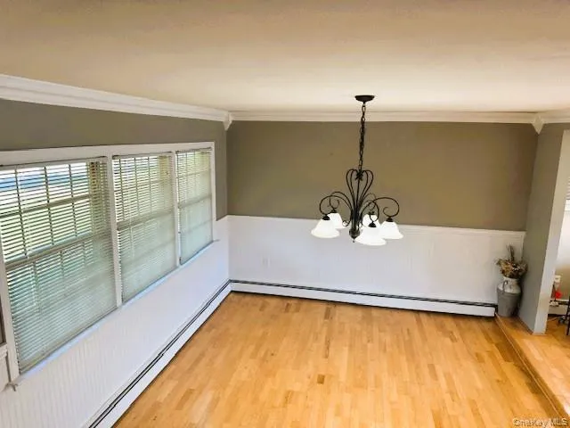 Unfurnished dining area featuring a baseboard radiator, a chandelier, a wainscoted wall, light wood-type flooring, and crown molding Unfurnished dining area featuring a baseboard radiator, a chandelier, a wainscoted wall, light wood-type flooring, and crown molding