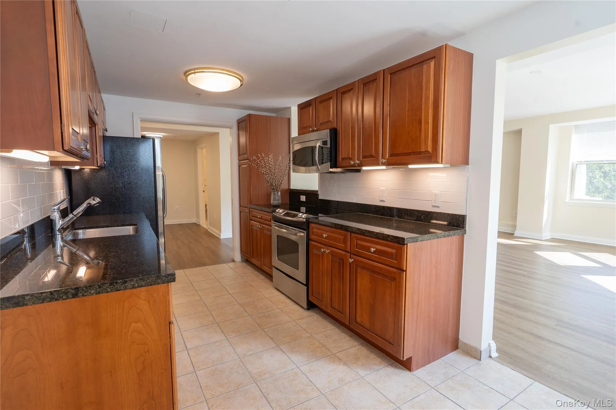 Kitchen featuring tasteful backsplash, wood cabinets, light tile patterned floors, dark stone counters, and stainless steel appliances; This view is looking into living room and front entrance simultanously Kitchen featuring tasteful backsplash, wood cabinets, light tile patterned floors, dark stone counters, and stainless steel appliances; This view is looking into living room and front entrance simultanously