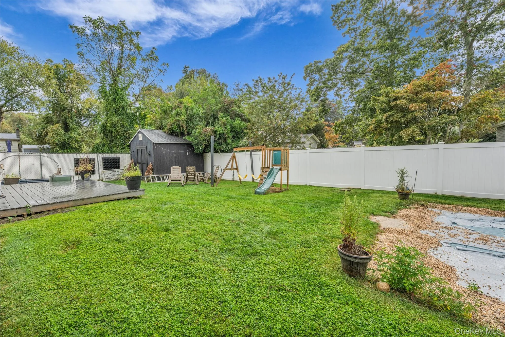 Fenced backyard with a playground, a storage unit, view of scattered trees, and a wooden deck Fenced backyard with a playground, a storage unit, view of scattered trees, and a wooden deck
